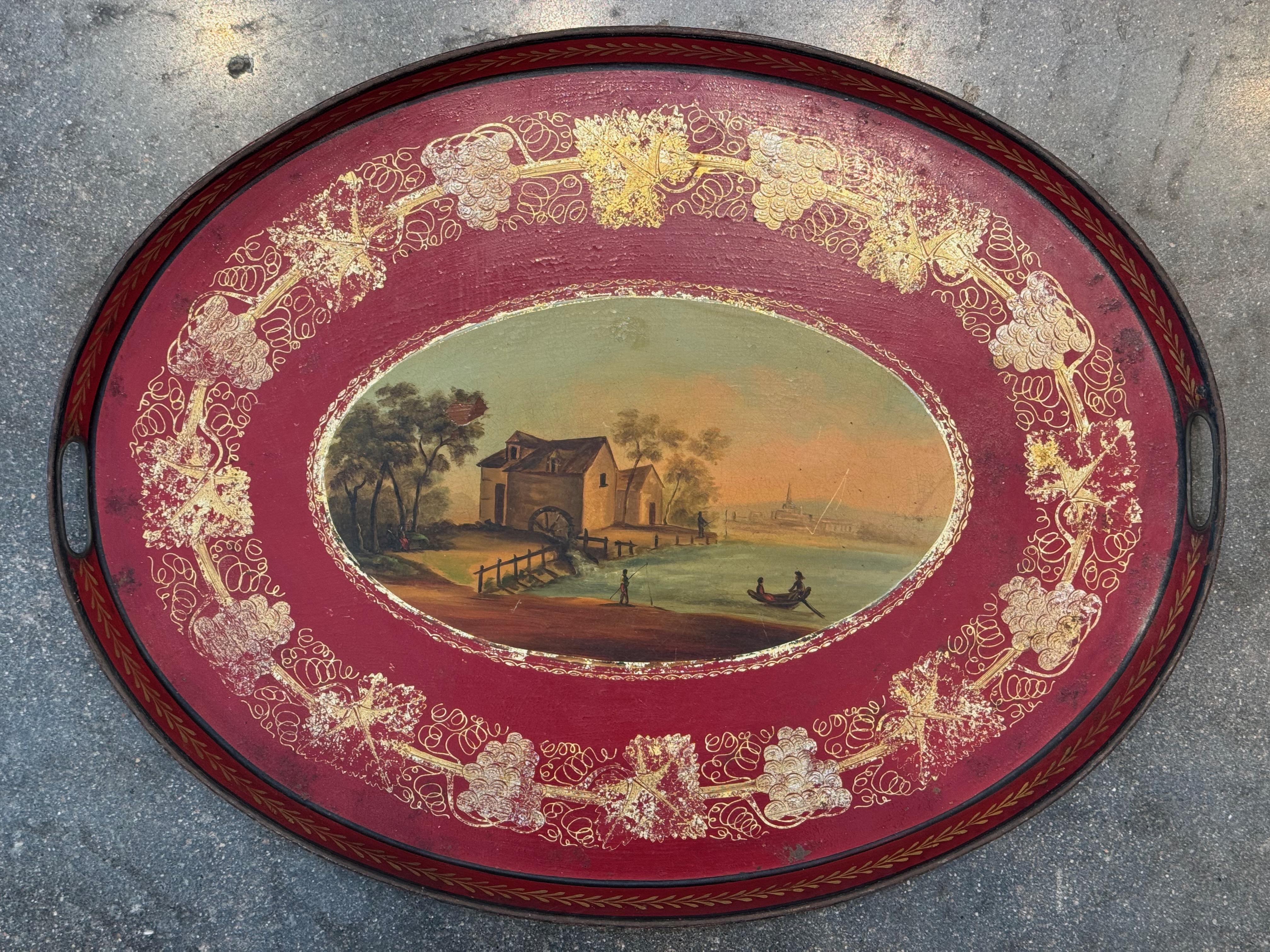 Vassoio ovale in tole francese del XIX secolo con una scena di porto riccamente dipinta al centro, delimitata da un fondo rosso intenso con intricati motivi dorati di viti e uva. I manici forati e il bordo nero completano lo stile classico del