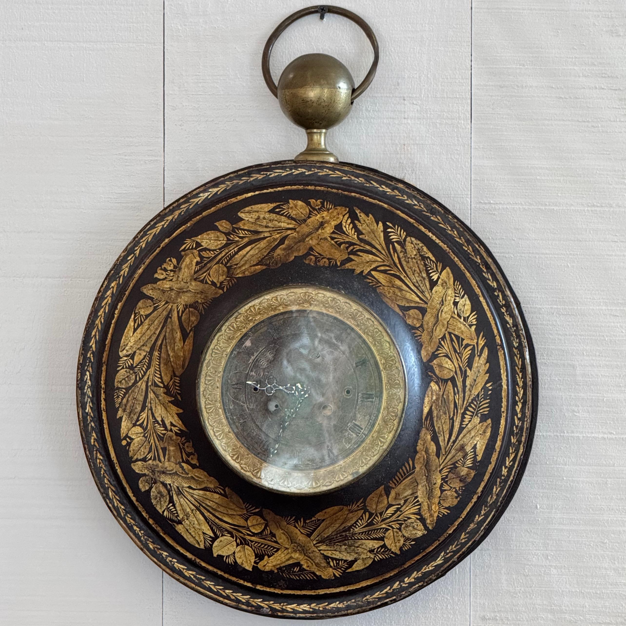 Decorated with finely detailed gilt foliate and bird motifs, this 19th century French tôle wall clock features a domed face with Roman numerals and ornate hands. The black lacquered surface is contrasted by rich gold accents and a brass ring finial