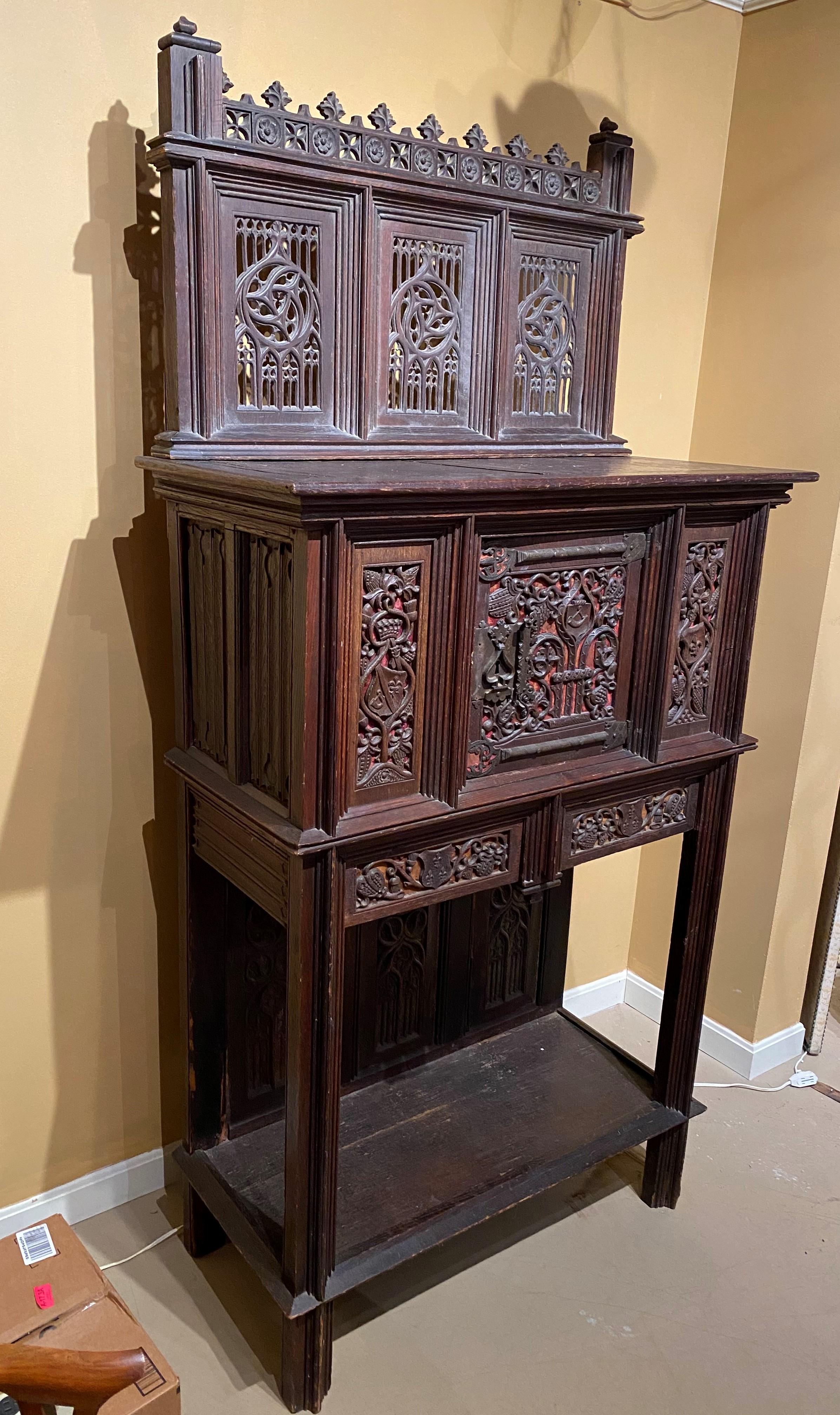 A fine form two-piece oak Gothic style cabinet in old dark finish with trefoil lined crest surmounting an upper tracery gallery back, over a single door cabinet, the center panel adorned with large decorative metal hinges and reticulated carved