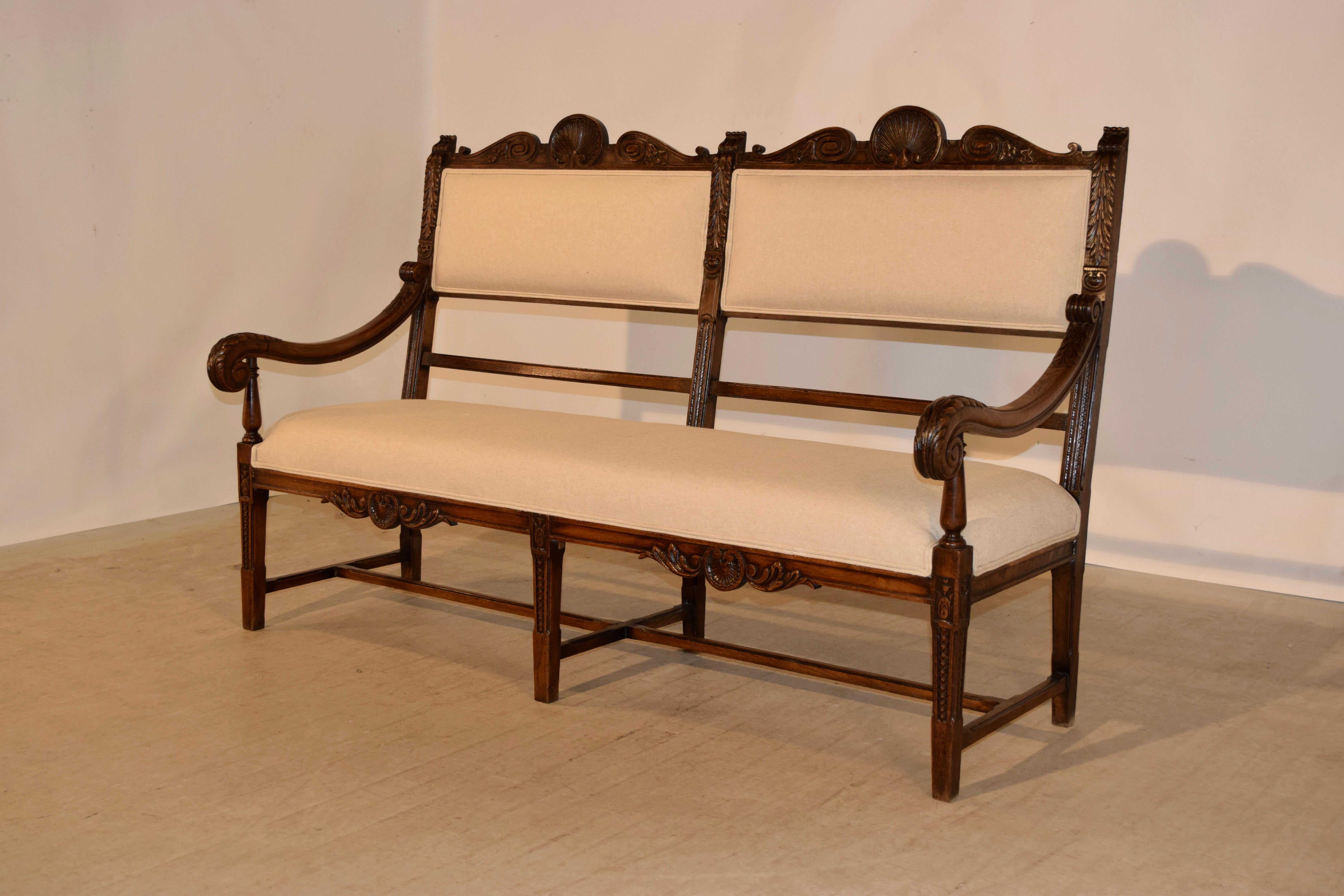 19th century upholstered bench from France made from oak with newly upholstered seat and back. The frame is ornately hand carved and has shell and leaf patterns with hand carved scrolled arms and supported on hand-turned legs joined by simple