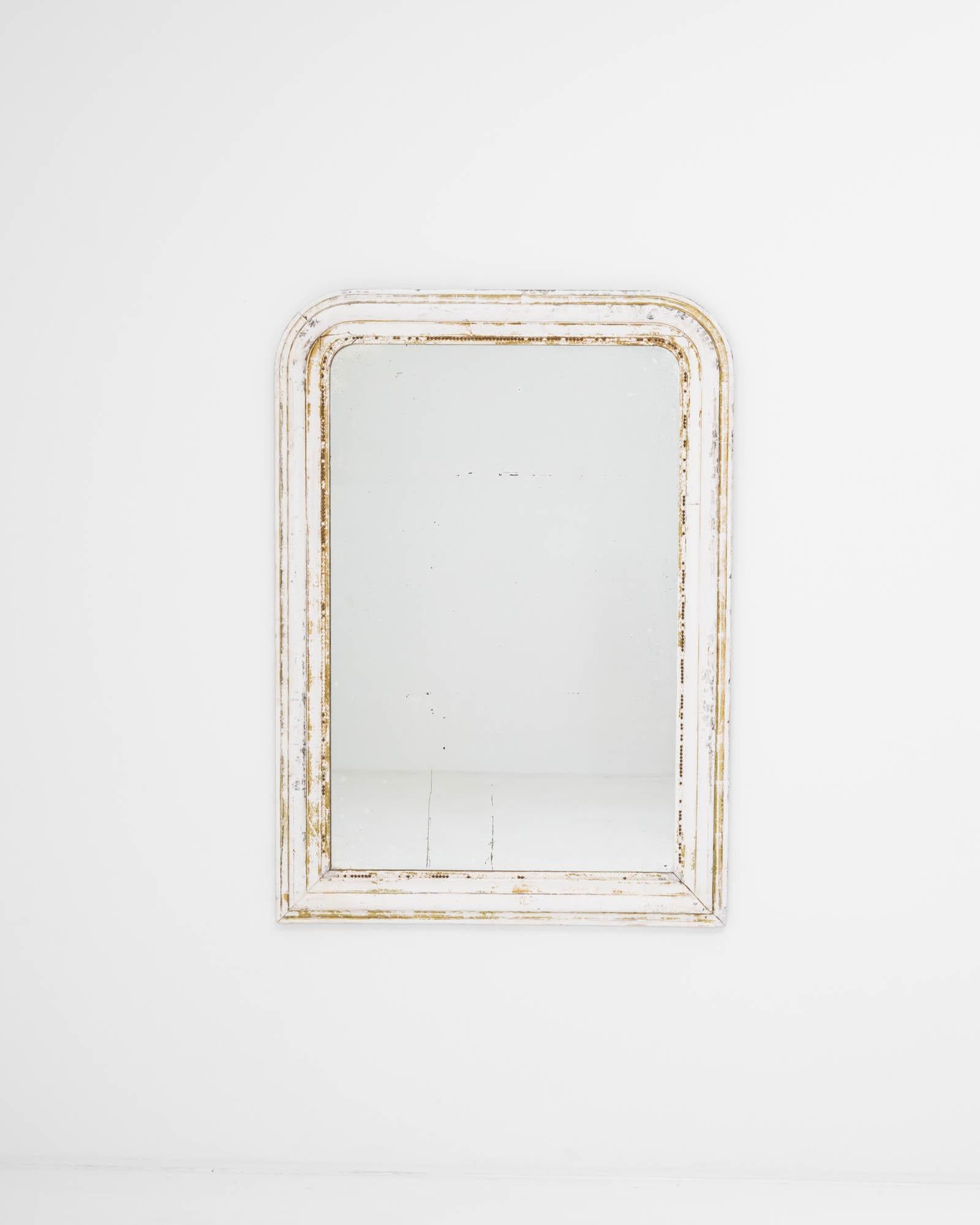 A wooden mirror created in 19th century France. Proudly aged, the white paint has worn to a gentle patina, unveiling warm hues that shine in accord with golden detailing that rings the arched frame. This one of a kind mirror combines traditional