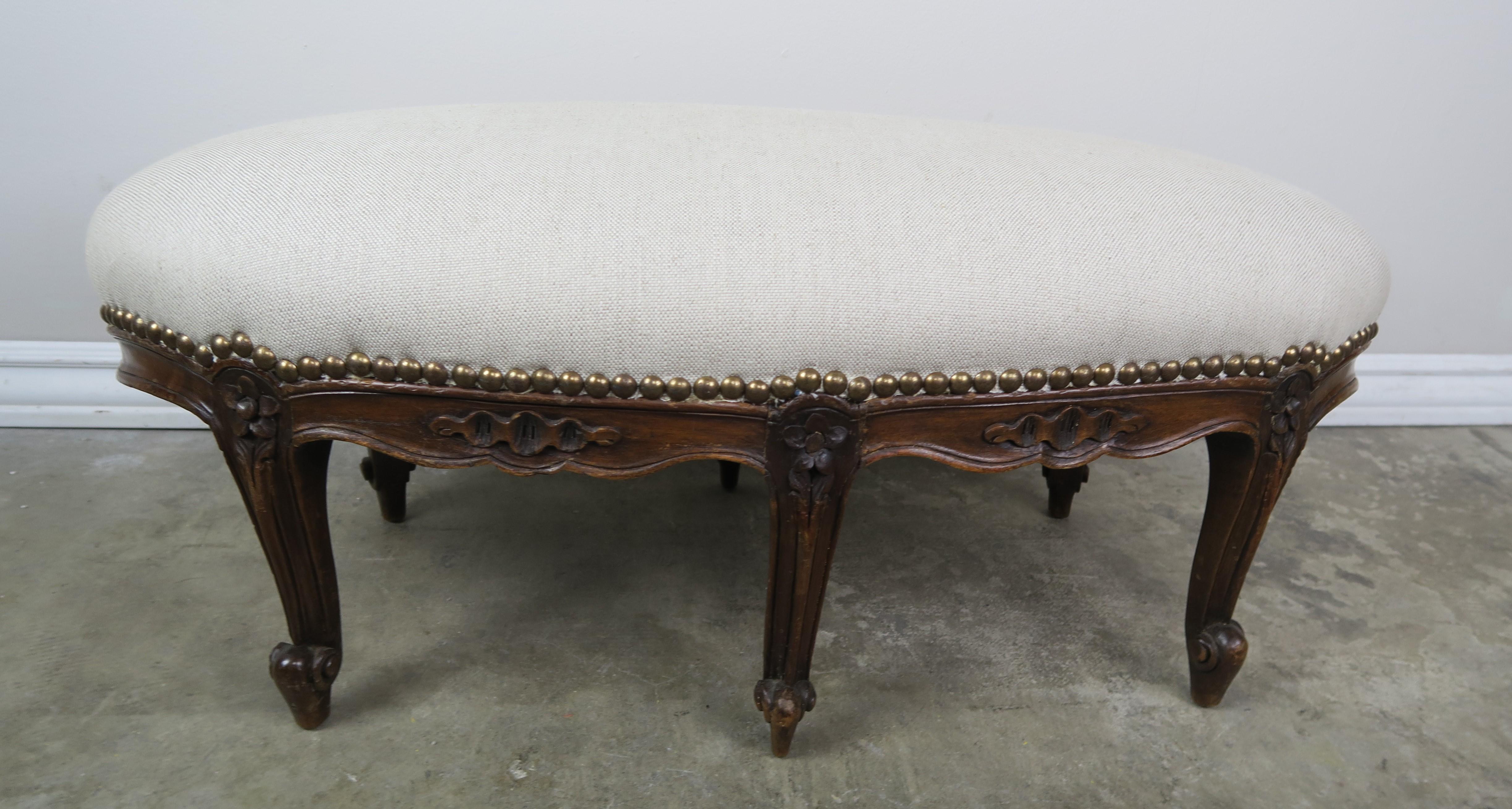 19th century oval shaped French Louis XV style walnut footstool standing on six cabriole legs ending in rams head feet. The footstool is newly upholstered in a Belgium linen with antique brass nailhead trim detail.