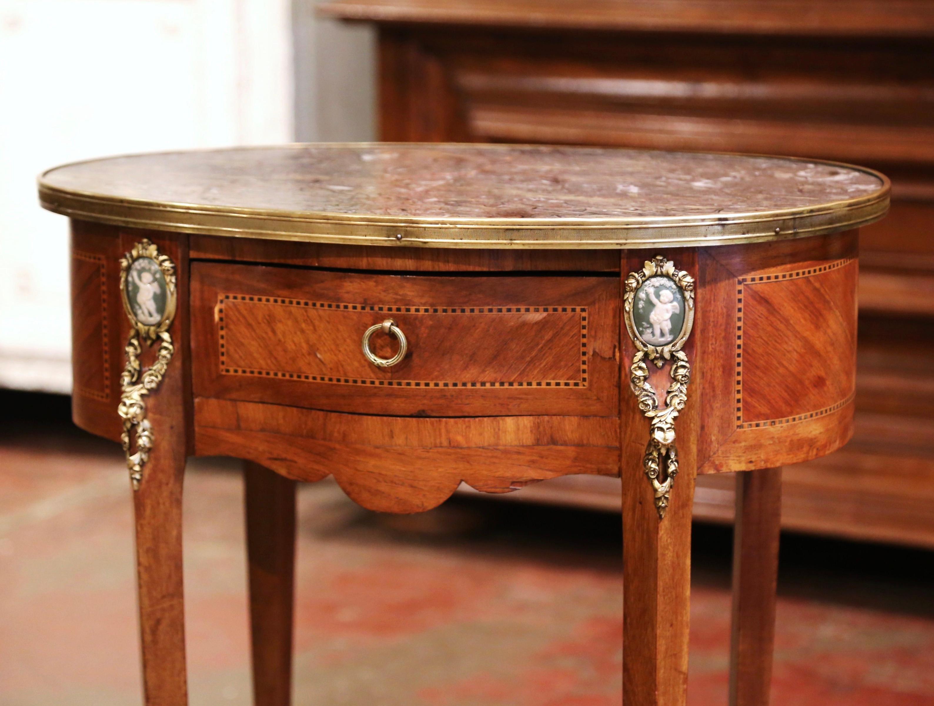 Crafted in France circa 1880, the elegant antique table stands on cabriole legs decorated with cherub porcelain plaque mounts at the shoulder, and ending with sabot feet. The table features detailed inlaid marquetry work around the apron, and is