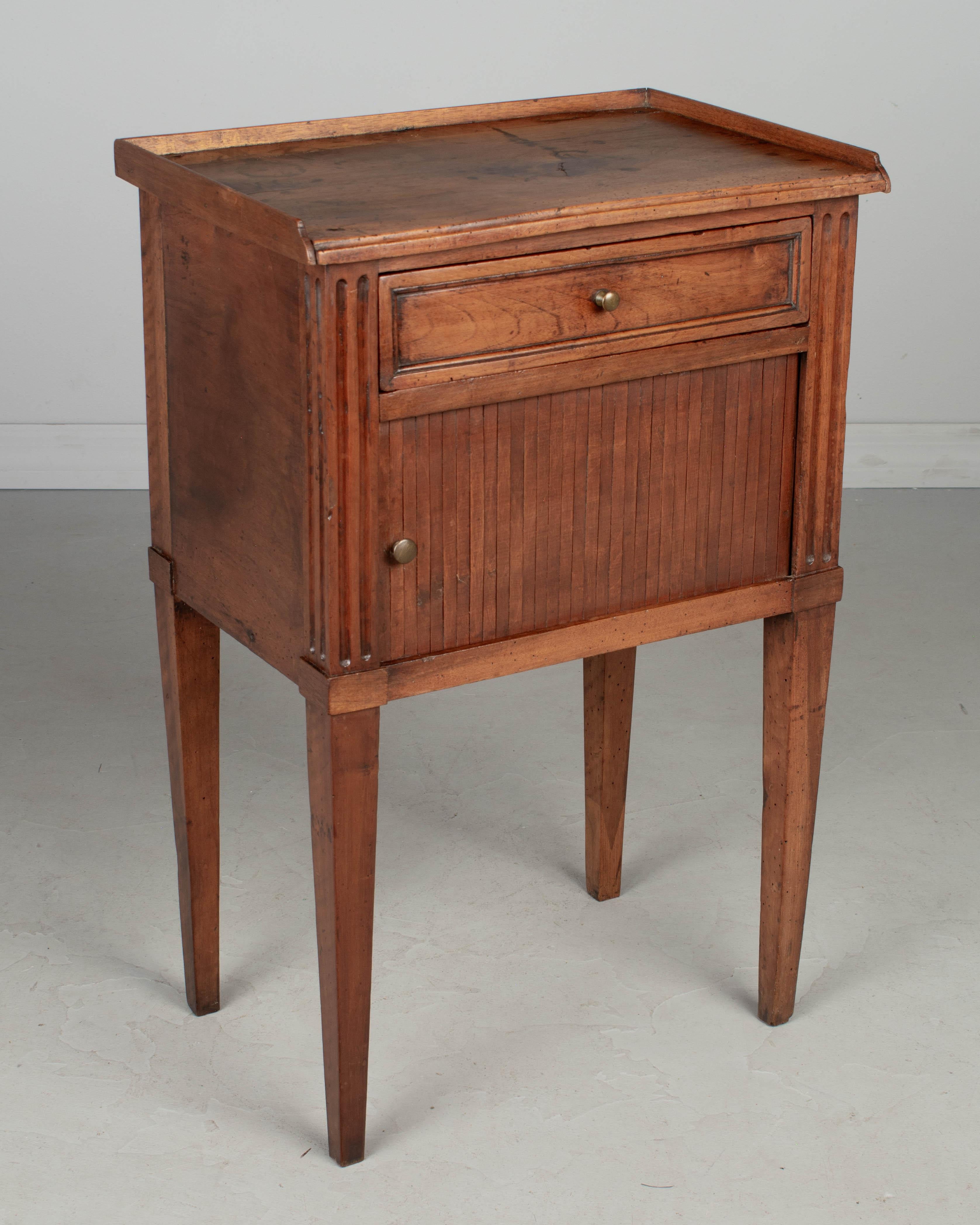 A 19th century French Country walnut side table or nightstand with a tambour door, dovetailed drawer and tapered legs. Brass knobs. Waxed patina. All original. Back right leg restored. Circa 1860-1880.   18.25