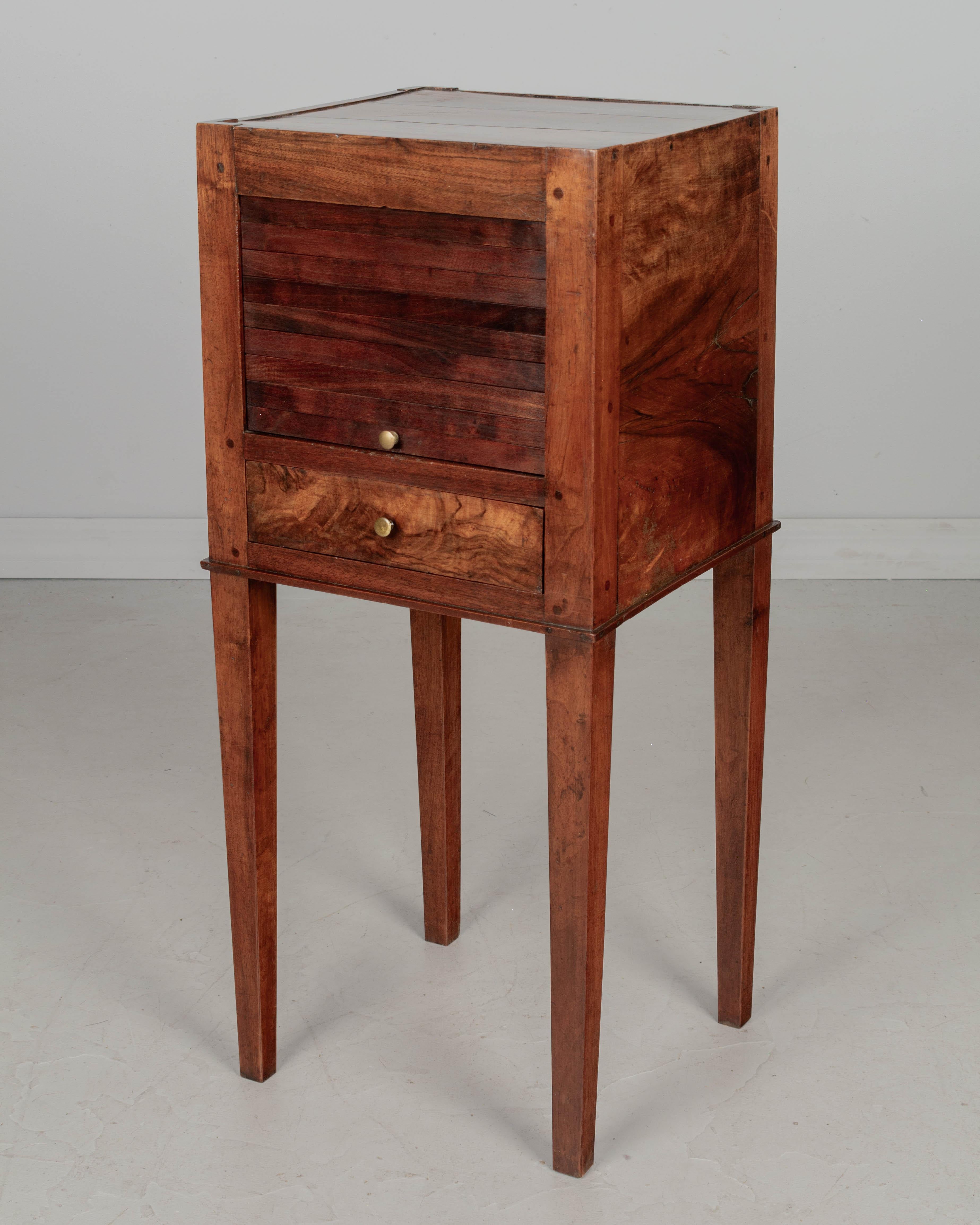 A 19th Century French walnut side table with a vertical tambour door and dovetailed drawer with brass knob. Finished on all four sides. Slender tapered legs. Nice choice of wood with beautiful grain, bookmatched sides. Waxed patina. Pegged