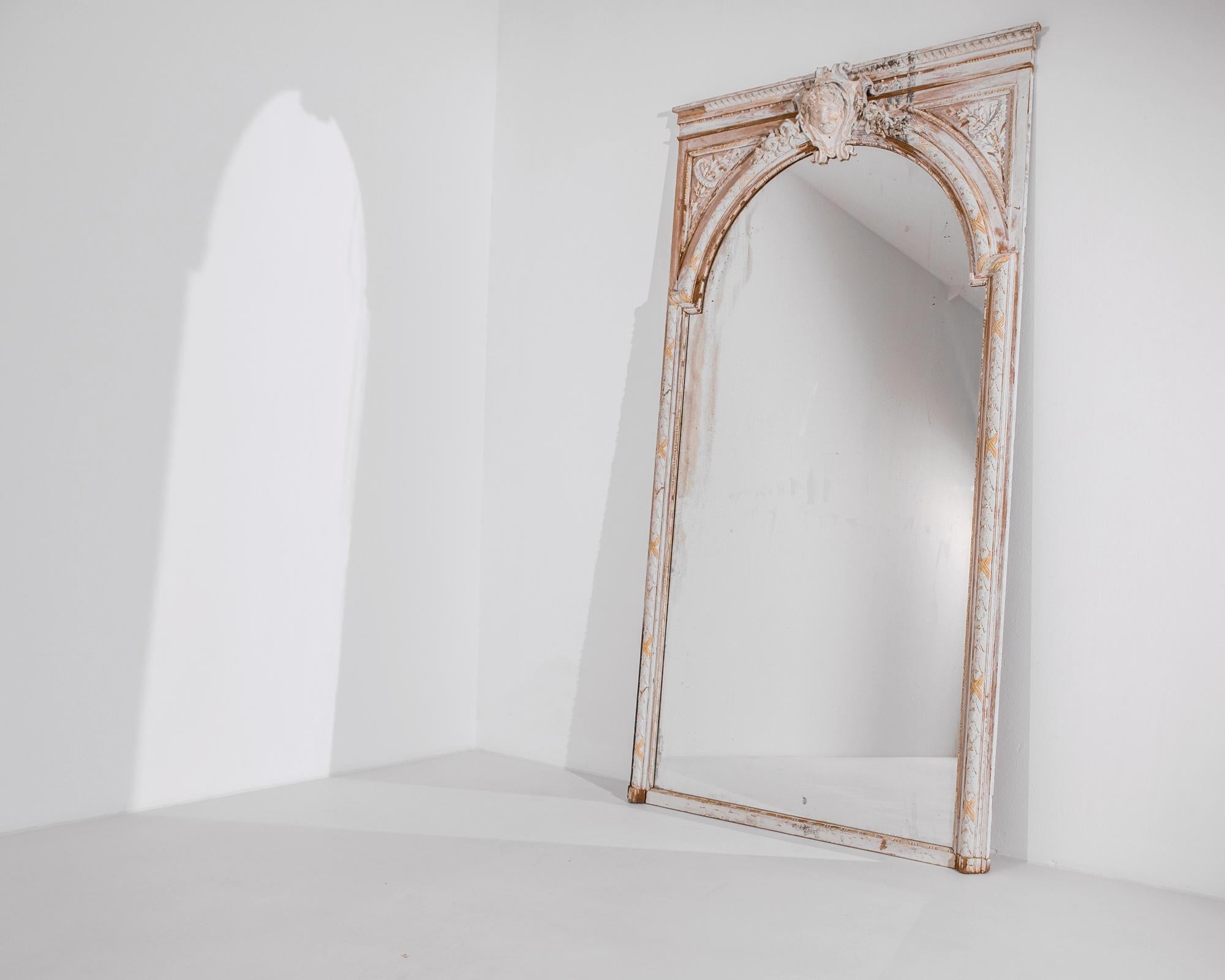 This antique wall mirror from France, circa 1860, offers an ornate carved frame gilded and adorned with a boyish mythological figure. An airy entryway expanding your interior space, the patinated gold and white paint gives this arched mirror a