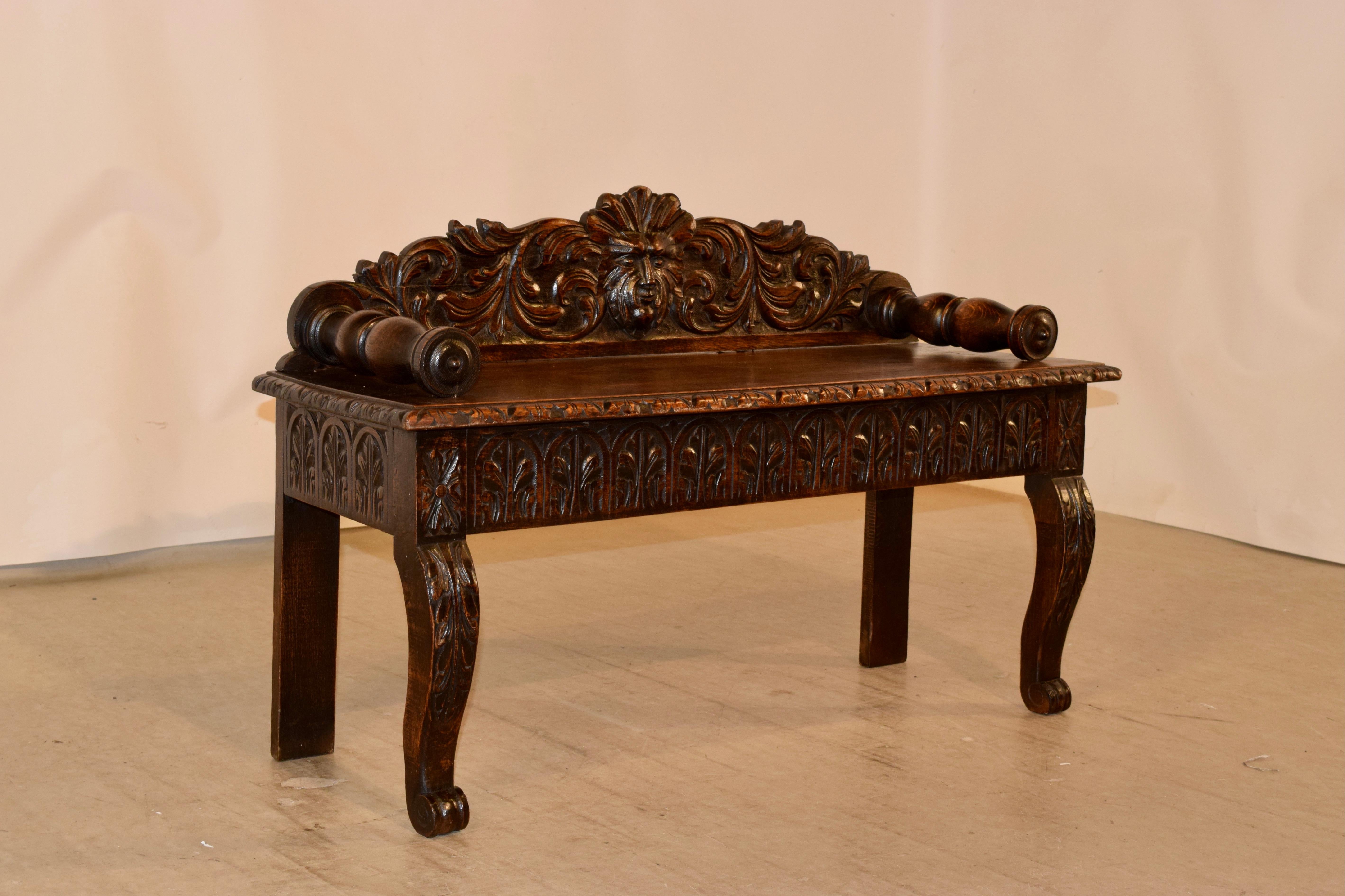 19th century oak window seat from France with an exquisitely hand carved back, depicting a central carved face surrounded by acanthus leaves. The arms are hand turned and are attached to the seat, which has a beveled and carved decorated edge,