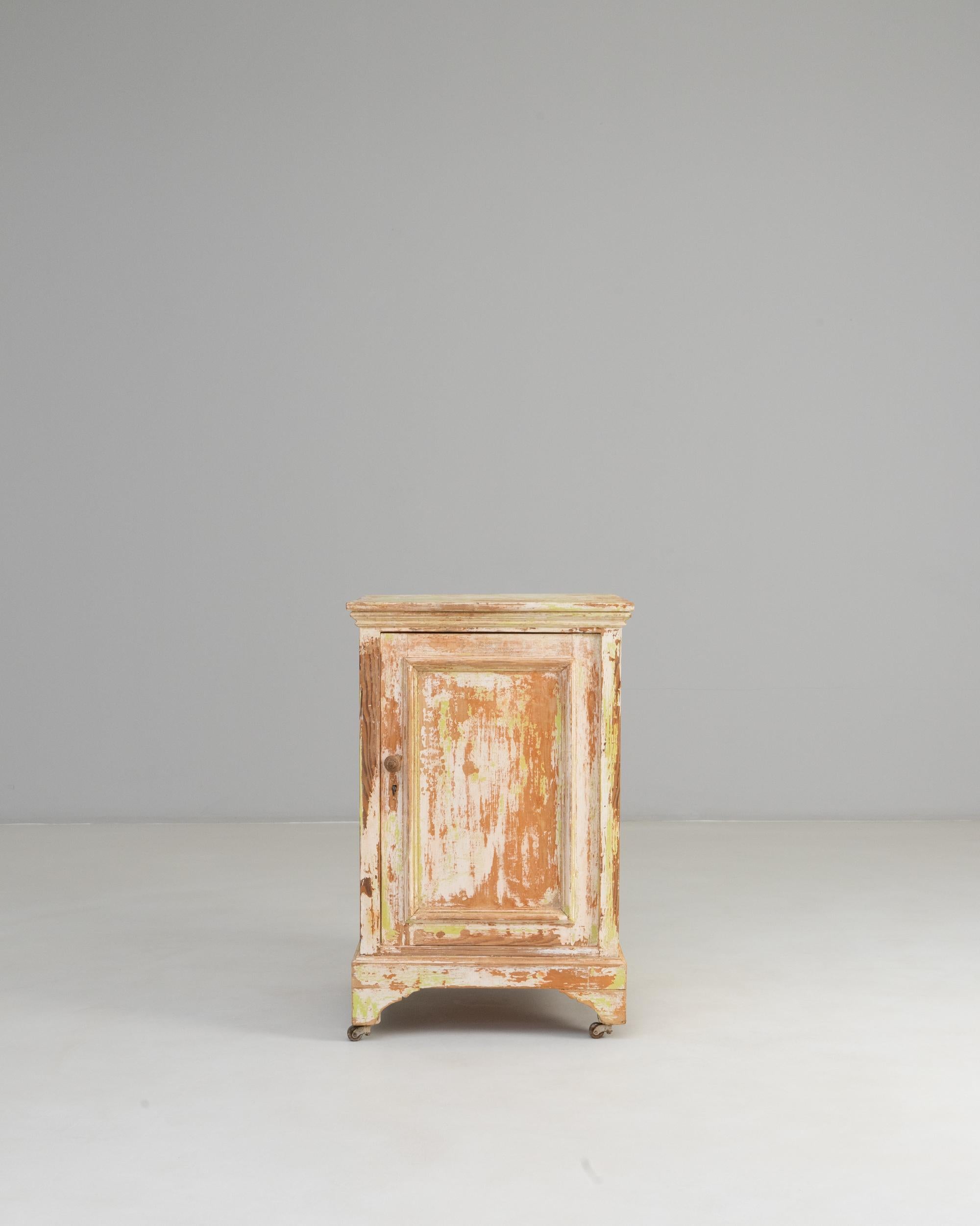 19th Century French Wood Patinated Cabinet With Wheels. This character-rich cabinet showcases the beautifully aged patina of its original painted finish, revealing layers of soft peach, cream, and hints of green that speak to decades of use and