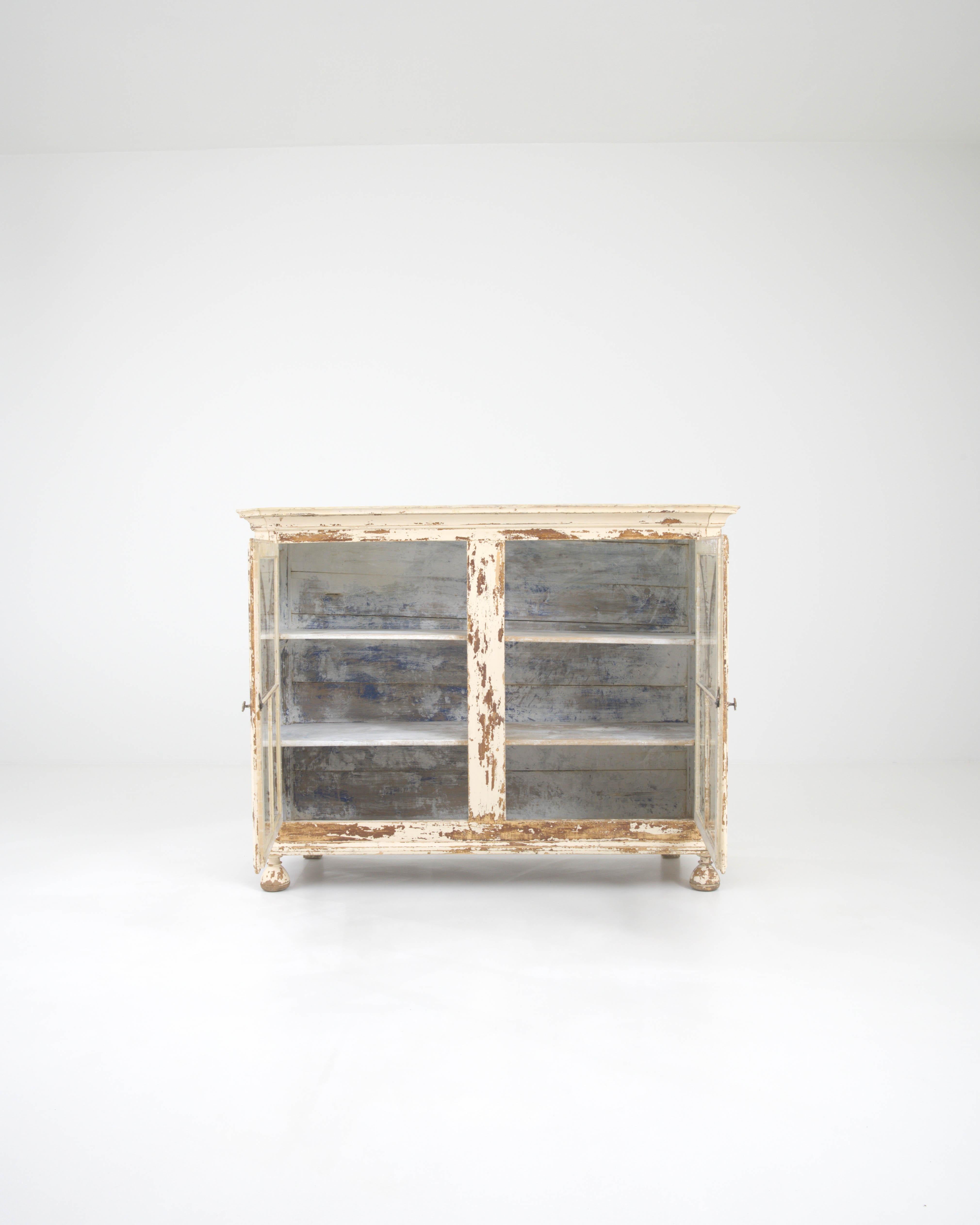 This 19th-century French wood vitrine captures the romantic essence of provincial life with its beautifully patinated finish and classic glass-paneled doors. The worn white paint reveals the time-weathered wood beneath, a testament to its journey