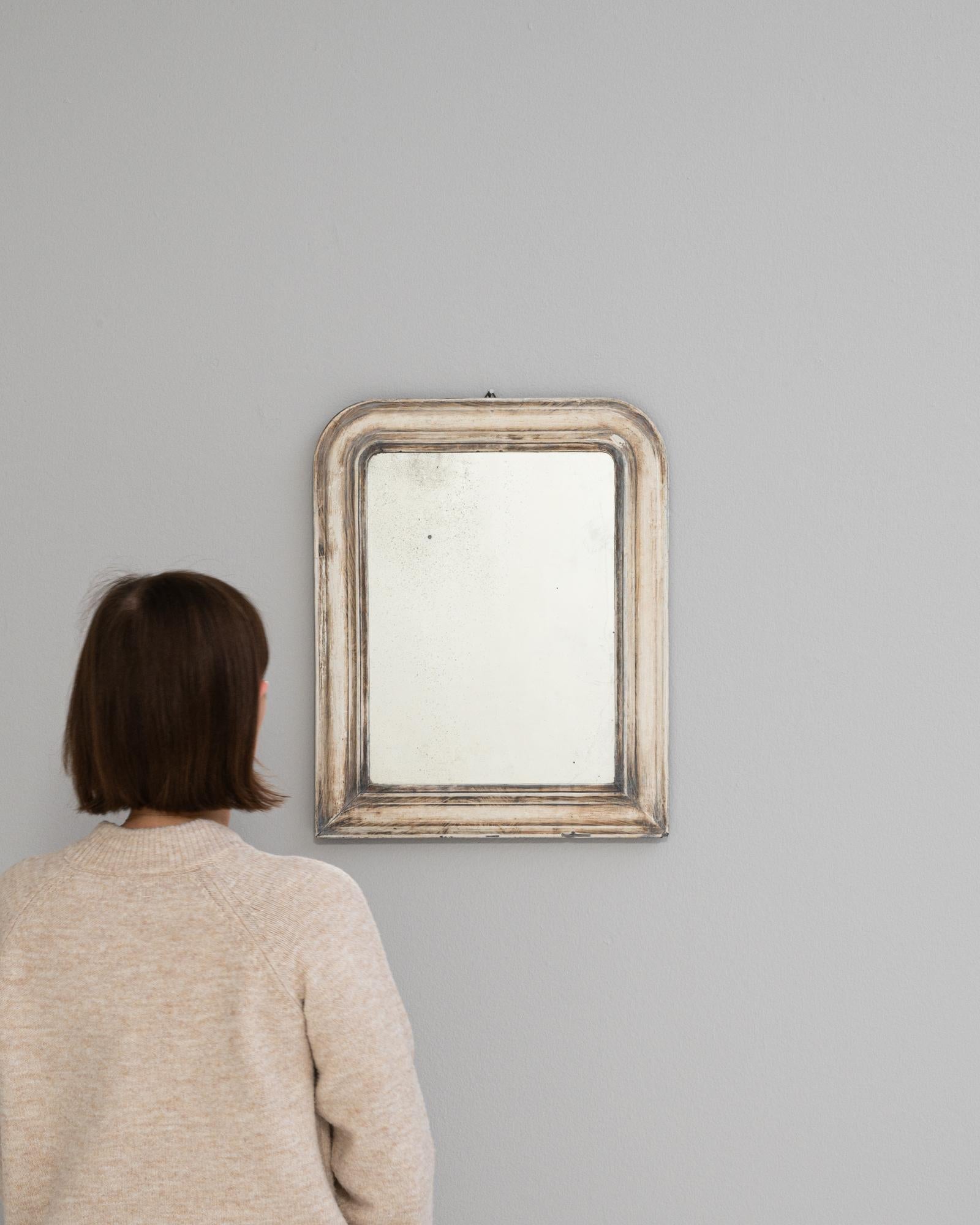 Discover the allure of antique charm with this 19th-century French wooden mirror, adorned with a white patina that tells a story of elegance and time. The gentle, rectangular frame, with its softly curved edges and weathered white finish, brings a