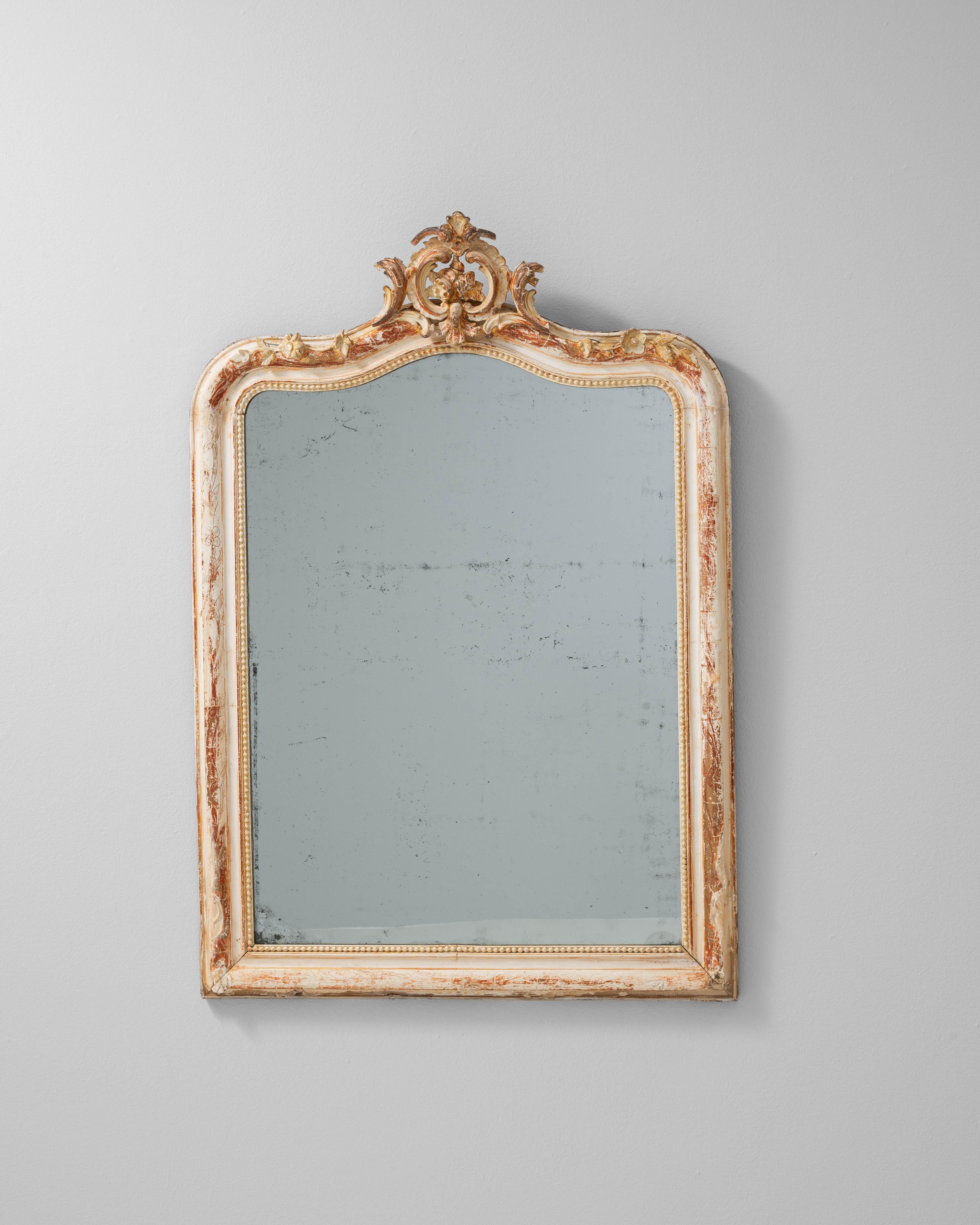 Este espejo francés del siglo XIX presenta un marco de madera elegantemente tallado, adornado con volutas y detalles florales de inspiración rococó. El acabado blanco patinado original, delicadamente desgastado con restos de dorado, realza su