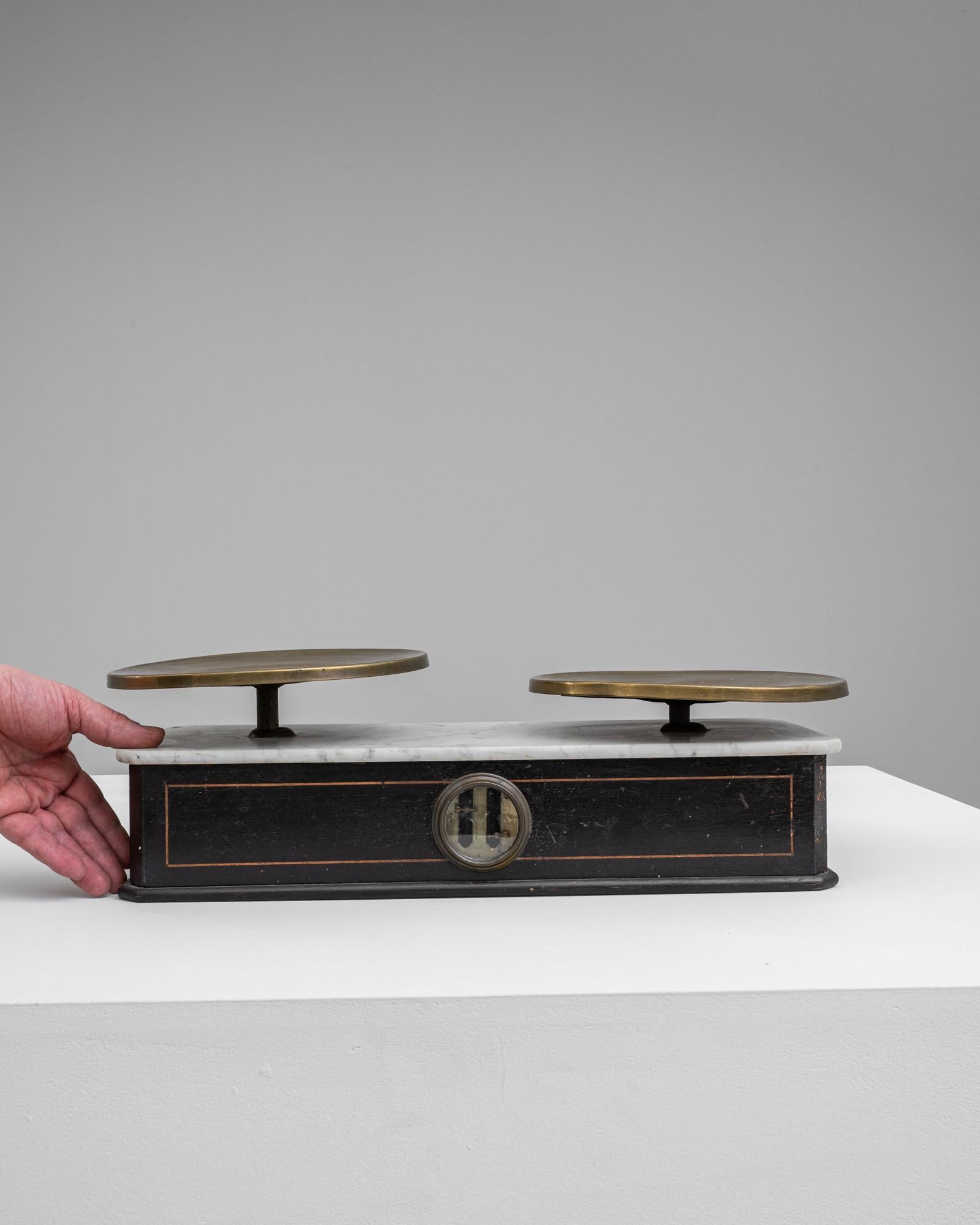 This 19th Century French Wooden & Metal Scale presents a stunning juxtaposition of elegance and precision. Its sleek wooden base, framed by delicate metal accents, provides a dignified stage for the polished brass pans that sit aloft. The central