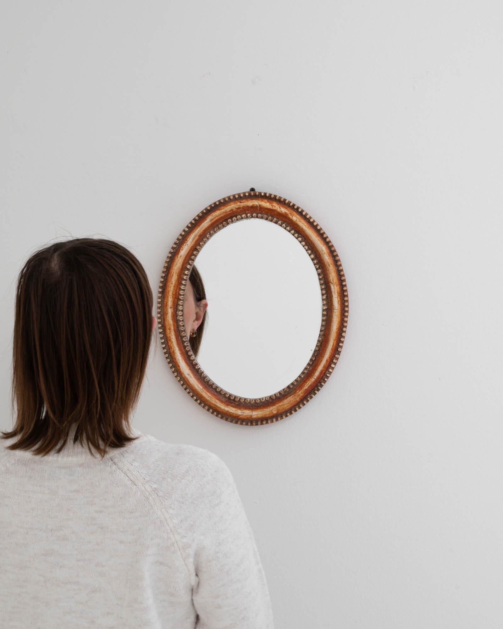 This 19th Century French wooden mirror is a charming artifact, exuding the elegance of a bygone era. The oval silhouette of the frame, adorned with intricate beading and subtle ornamentation, hints at the delicate craftsmanship of the period. The