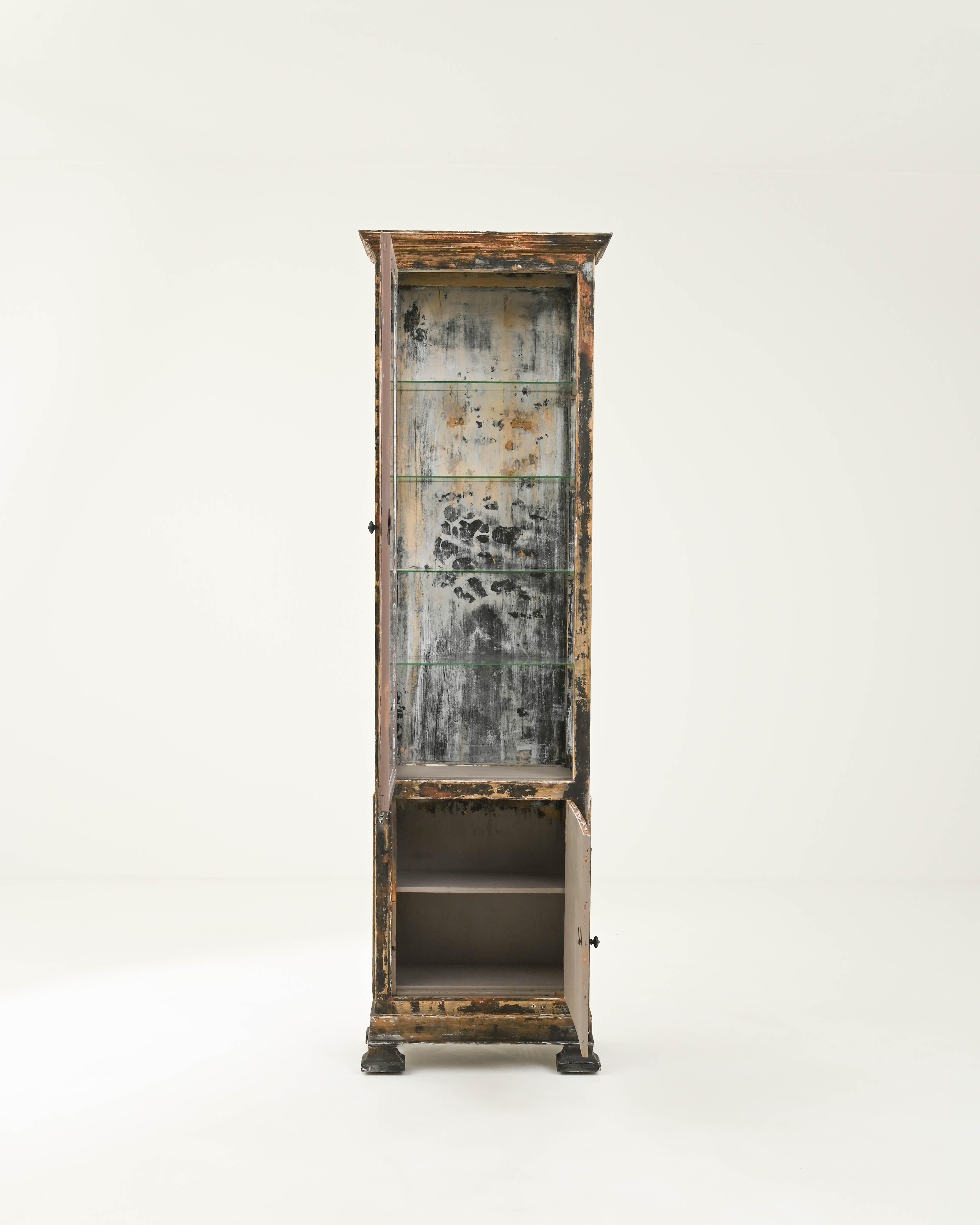 The tall, slender silhouette and dramatic patina of this wooden vitrine make it a covetable antique find. Built in France in the 1800s, windows on three sides of the narrow cabinet give a view onto four glass shelves, while a paneled cupboard below