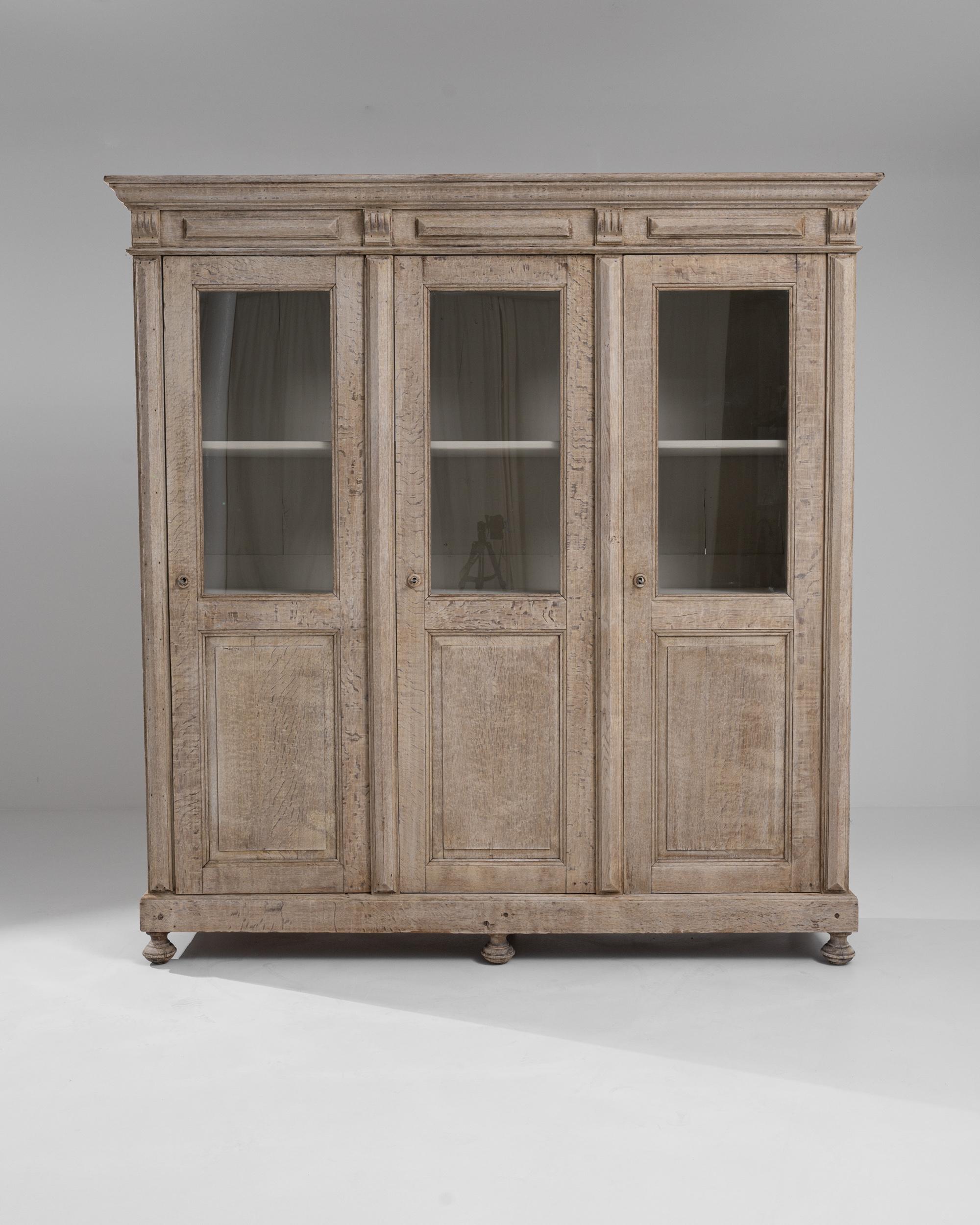 A bleached oak vitrine from 19th century France. Three paneled doors swing outward to reveal a wealth of interior storage, the top shelves on the display, while the bottom are concealed. The white, painted interior glows in synergy with the exterior