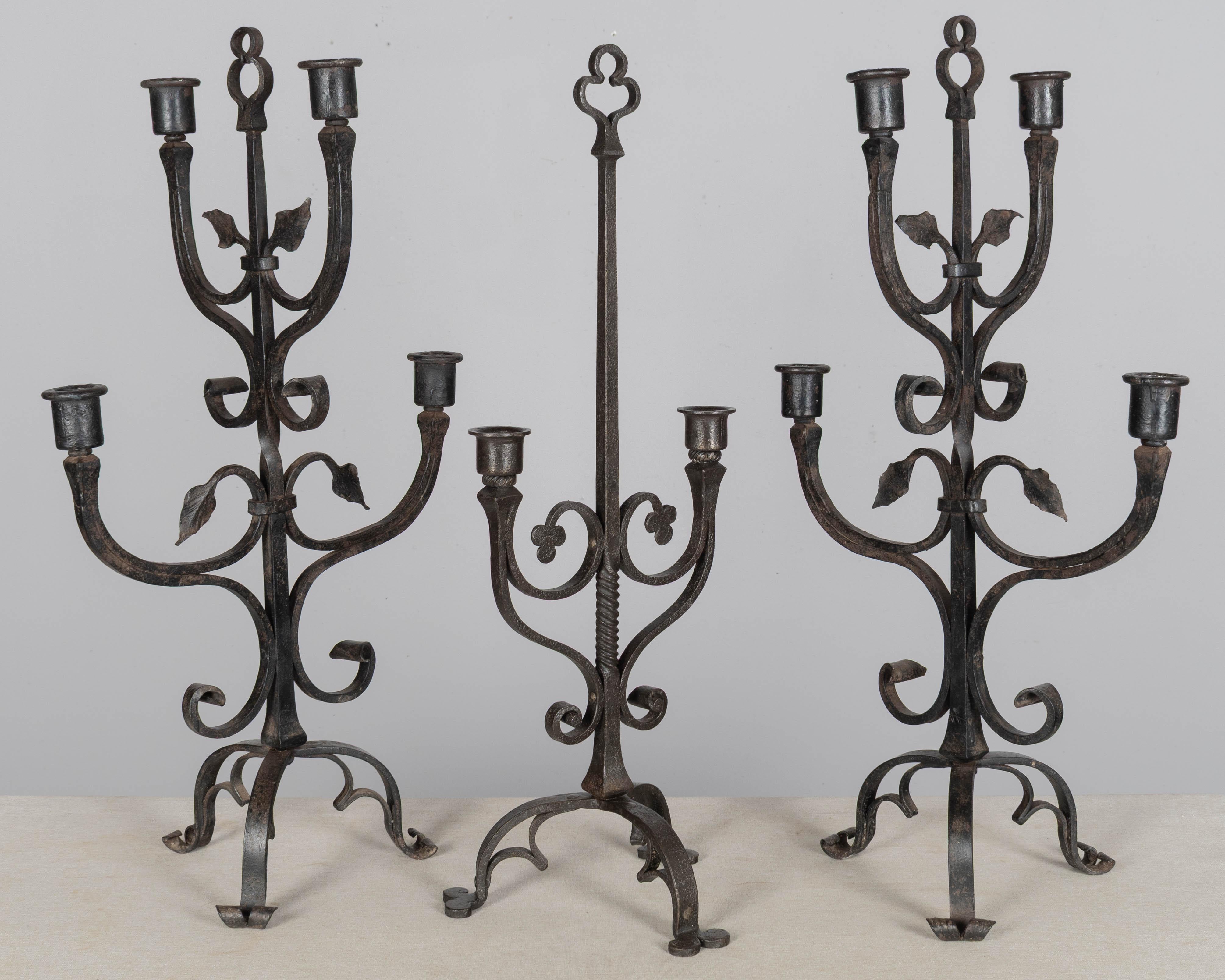 A set of three large French wrought iron candelabra with scroll form and rusty black patina. Good quality ironwork, riveted not welded. The pair has four candle cups and leaves and the single has two candle cups and twisted stem. Hand-forged with