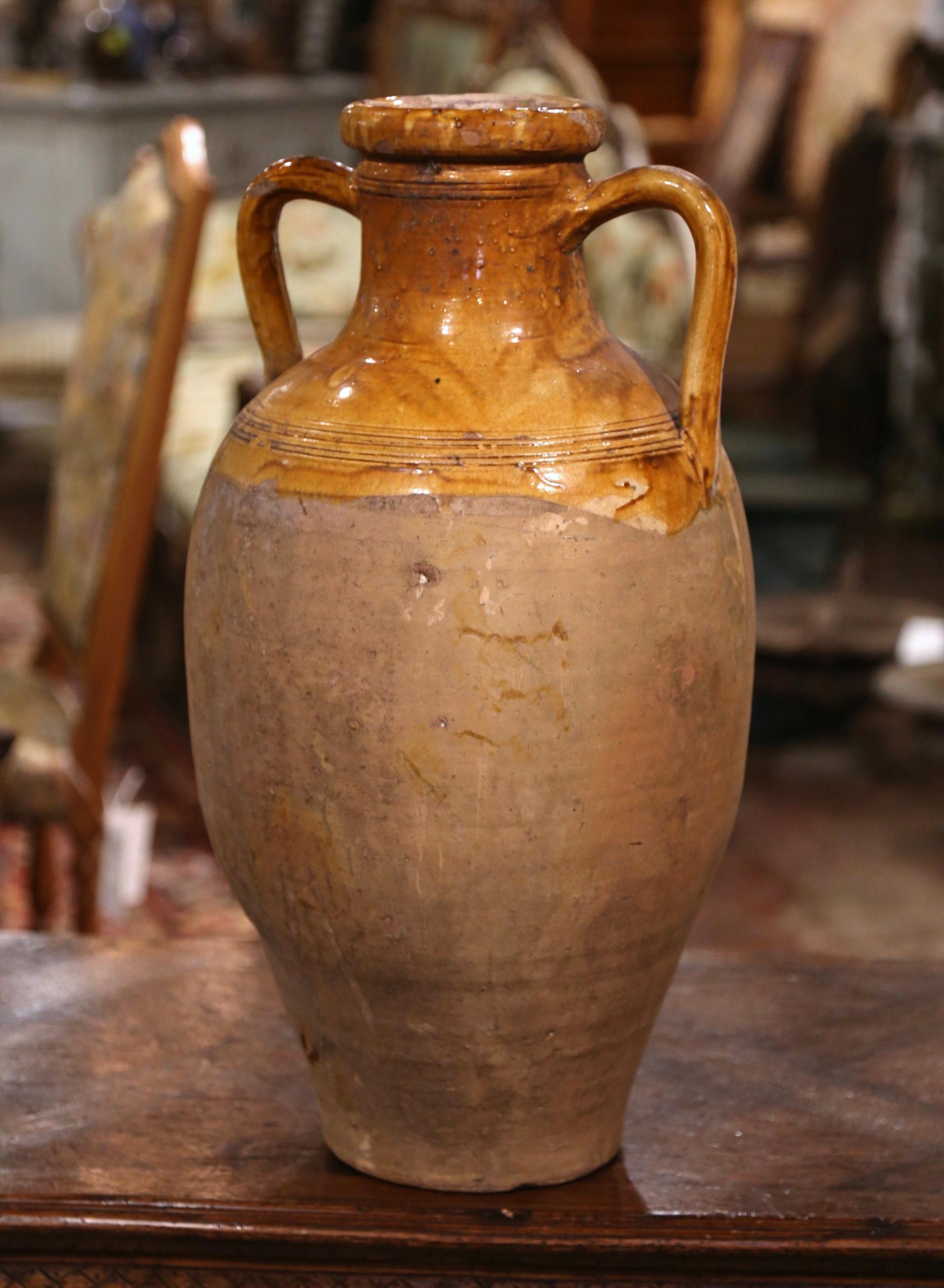 This large antique earthenware olive oil urn was created in Southern France, circa 1880. Made of blond clay and neutral in color, the terracotta vase has a traditional round shape decorated with side handles. The rustic, time-worn pot features a