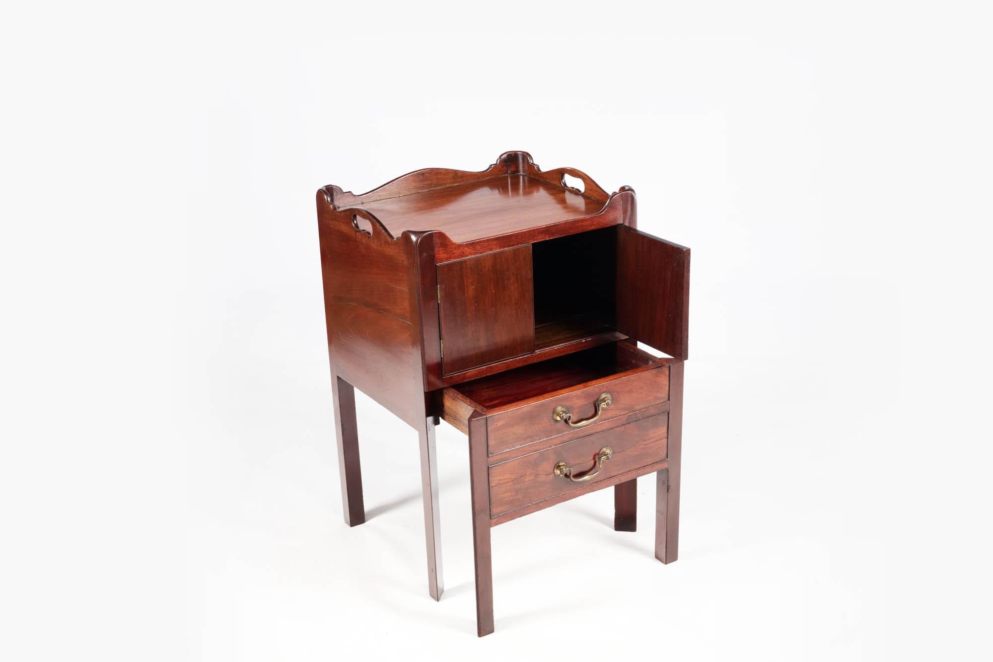 19th Century George III mahogany commode with tray top and brass handles. Of the late Georgian period, this mahogany commode features a scalloped-edge gallery top with pierced handles and two cupboard doors above a deep pull-out drawer, which was