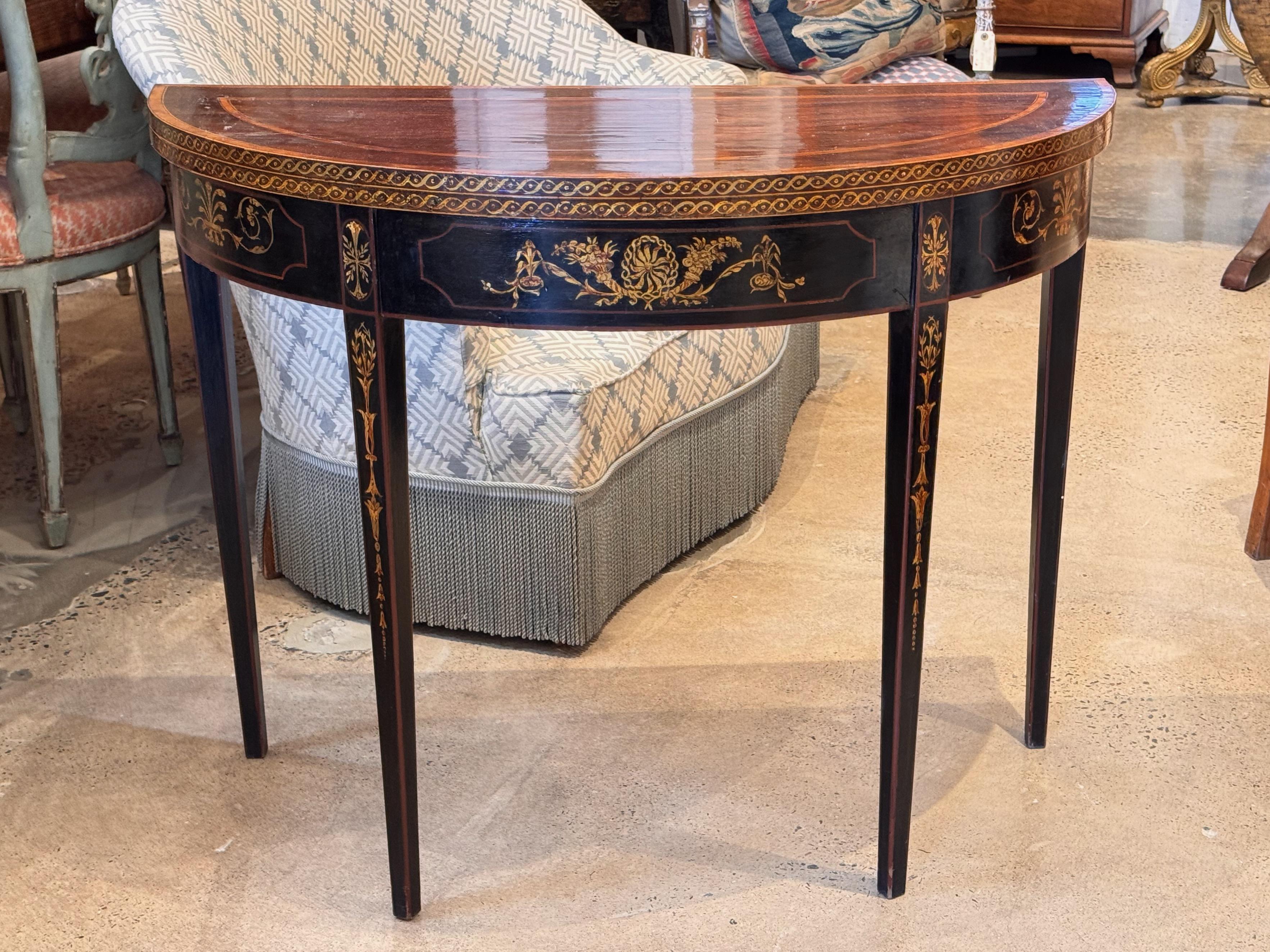 Elegant George III demilune card table with rich mahogany top and ebonized base, detailed with fine gilt and painted neoclassical motifs. The top folds open to reveal a green baize playing surface, supported by a swing-out rear leg. Graceful tapered