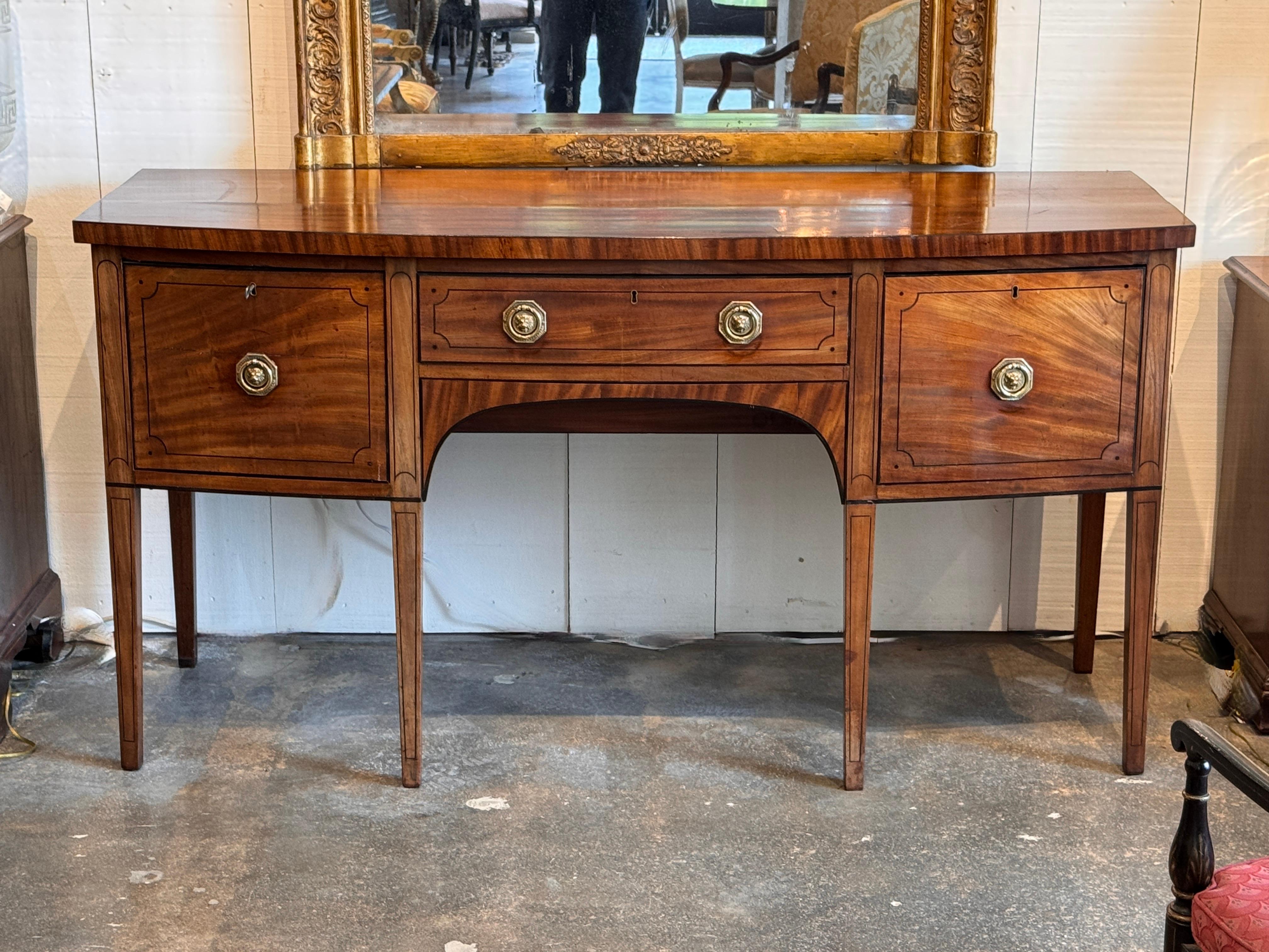 Mahogany bowfront sideboard finely detailed with satinwood and ebony stringing. The case features dovetailed drawers and original brass lion’s-head pulls, supported on square tapered legs. Rich figure to the wood and well-preserved craftsmanship