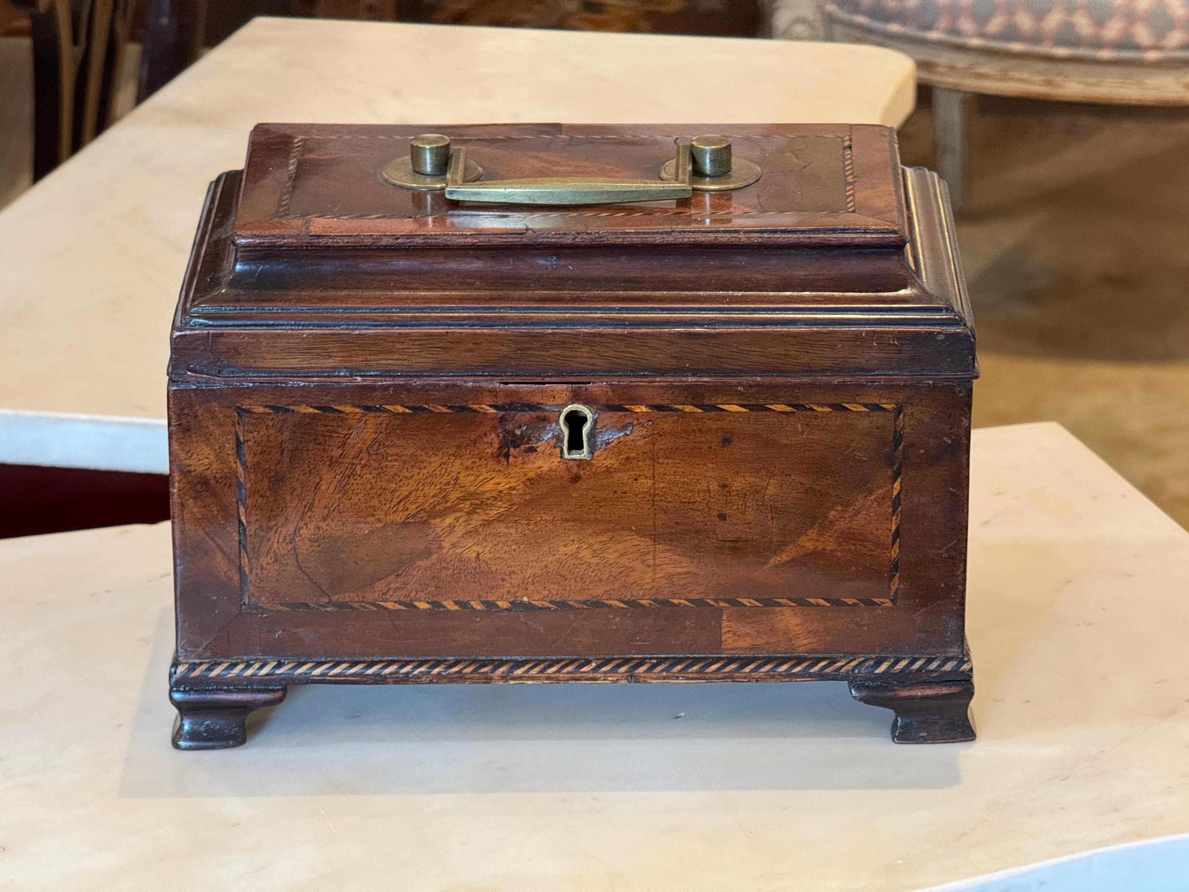 Early 19th century English tea caddy in the George III style, featuring finely inlaid panels and a brass swing handle on the raised lid. The interior is divided into three compartments with individual lids, all lined in green felt. The box sits on