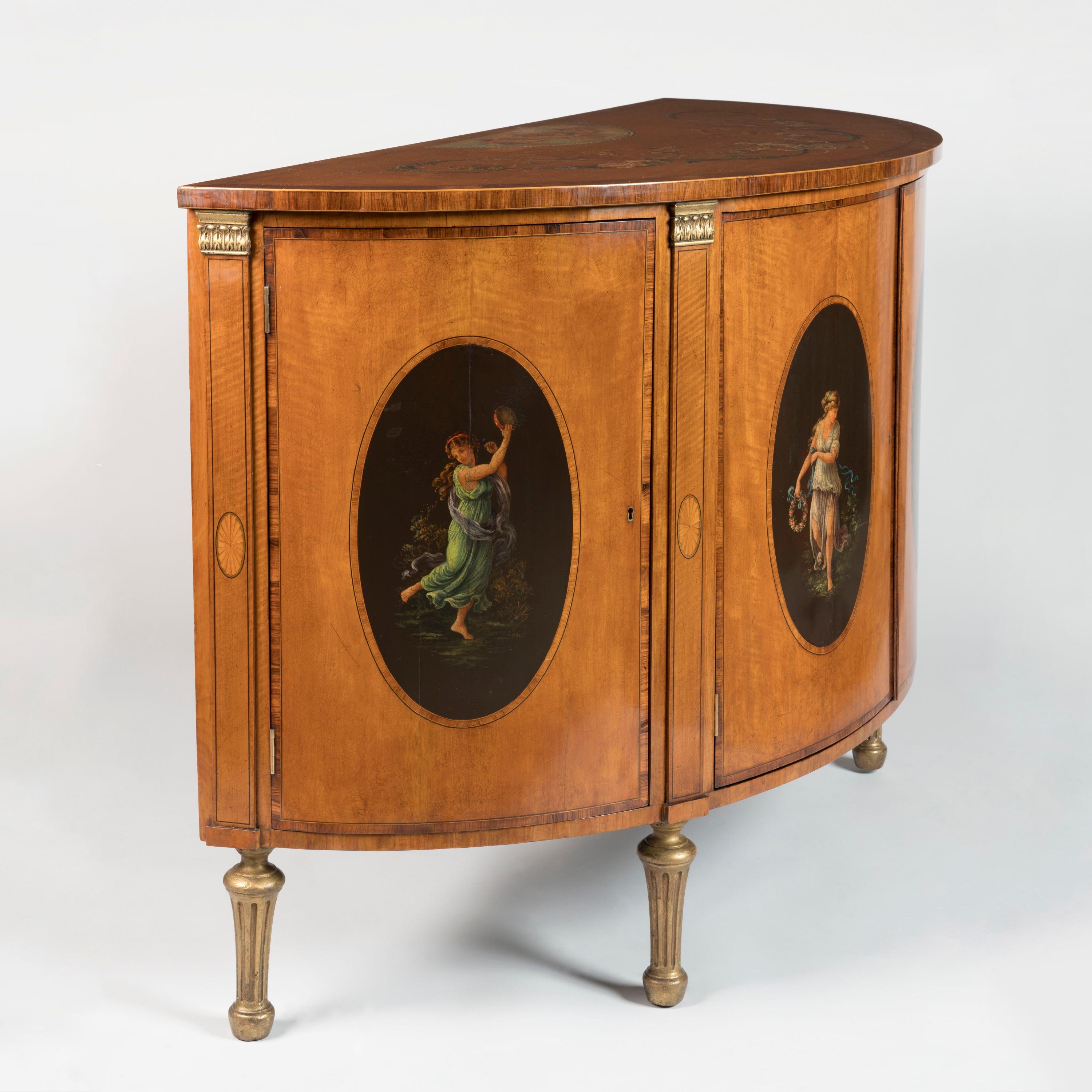 A Painted Satinwood Demilune Commode
In the George III Style

Constructed from satinwood and crossbanded with rosewood veneers, enlivened with boxwood stringing and giltwood details, the D-shaped top painted with a central scene of cherubs amidst