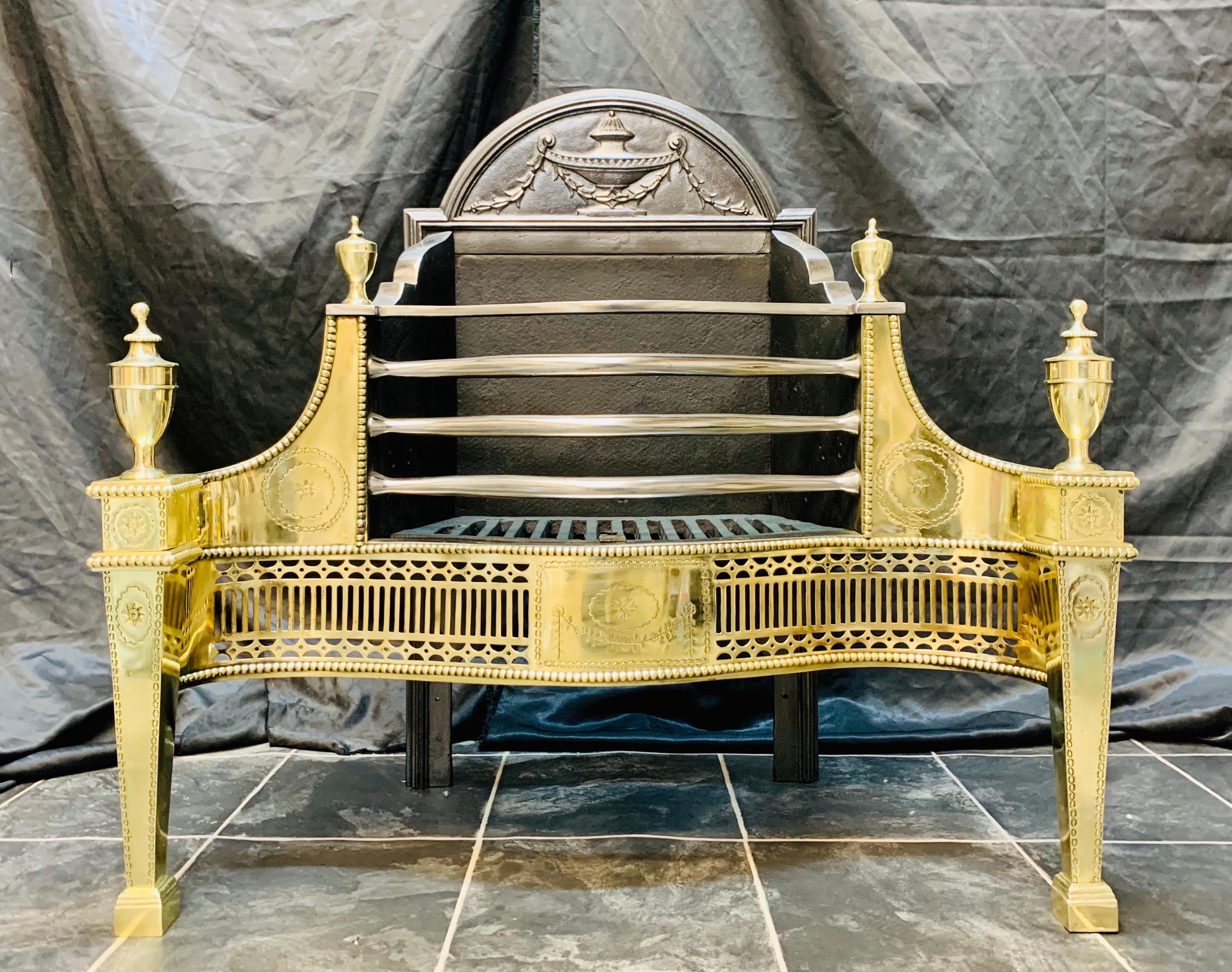 A Thomas Elsley, polished brass and cast iron Georgian style fire basket. The arched fireback centred by an urn draped with bellflowers above a four barred curved grate, with etched shoulders, is framed by beading and surmounted by a pair of lidded