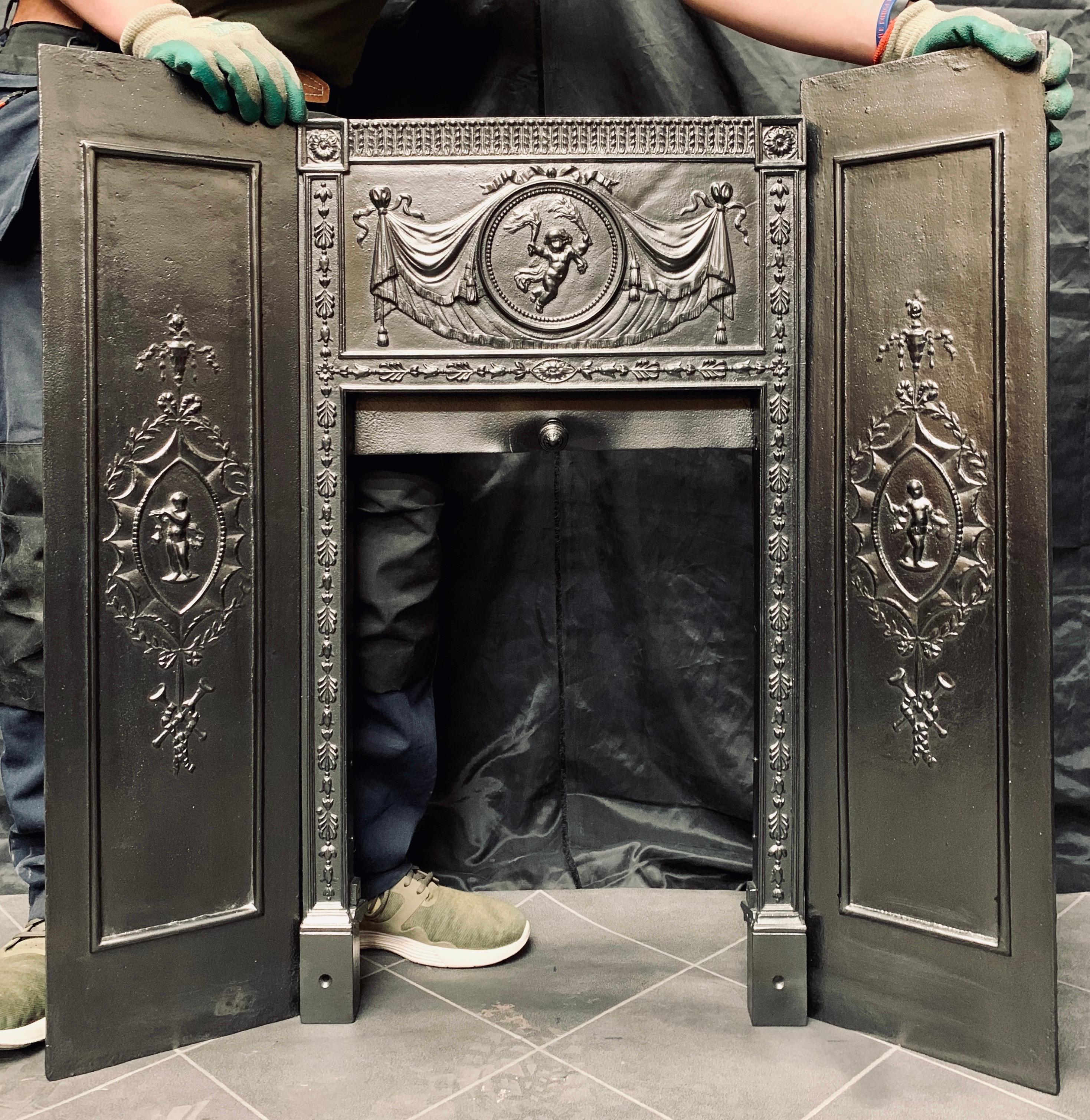 A rare 19th century Georgian Triptych paneled cast iron fireplace set. A central paneled grate opening with a central circular framed cherub holding flaming torches flanked by linen curtains and swags, below is a sliding door, the side panels are