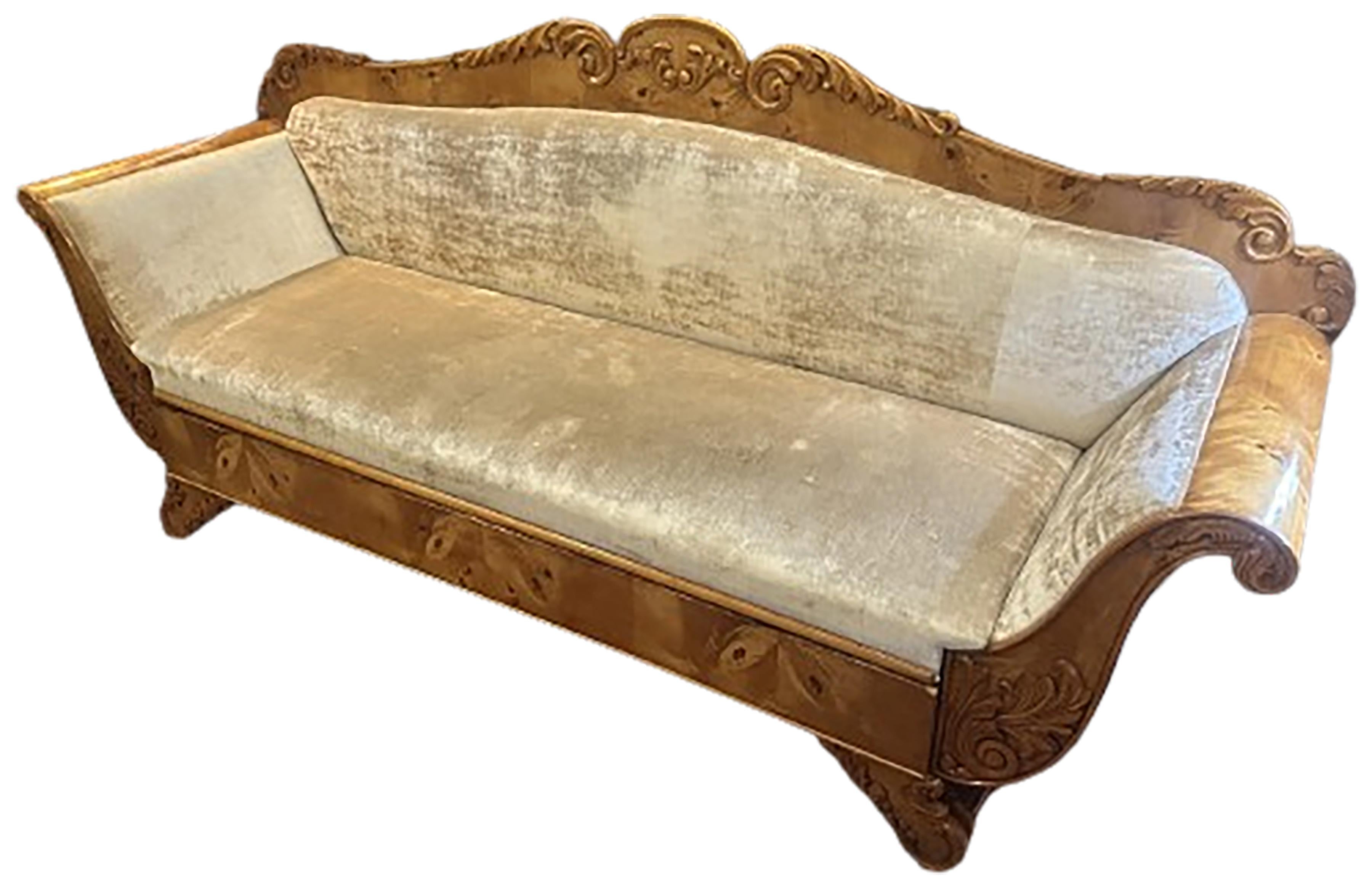 Un incantevole divano a due piazze Biedermeier. Struttura in legno di Wood con braccioli svasati e a voluta. Seduta, schienale e braccioli in velluto oro con cassetto per manubrio estraibile. Gambe e bracci intagliati a motivo di acanto. I cuscini