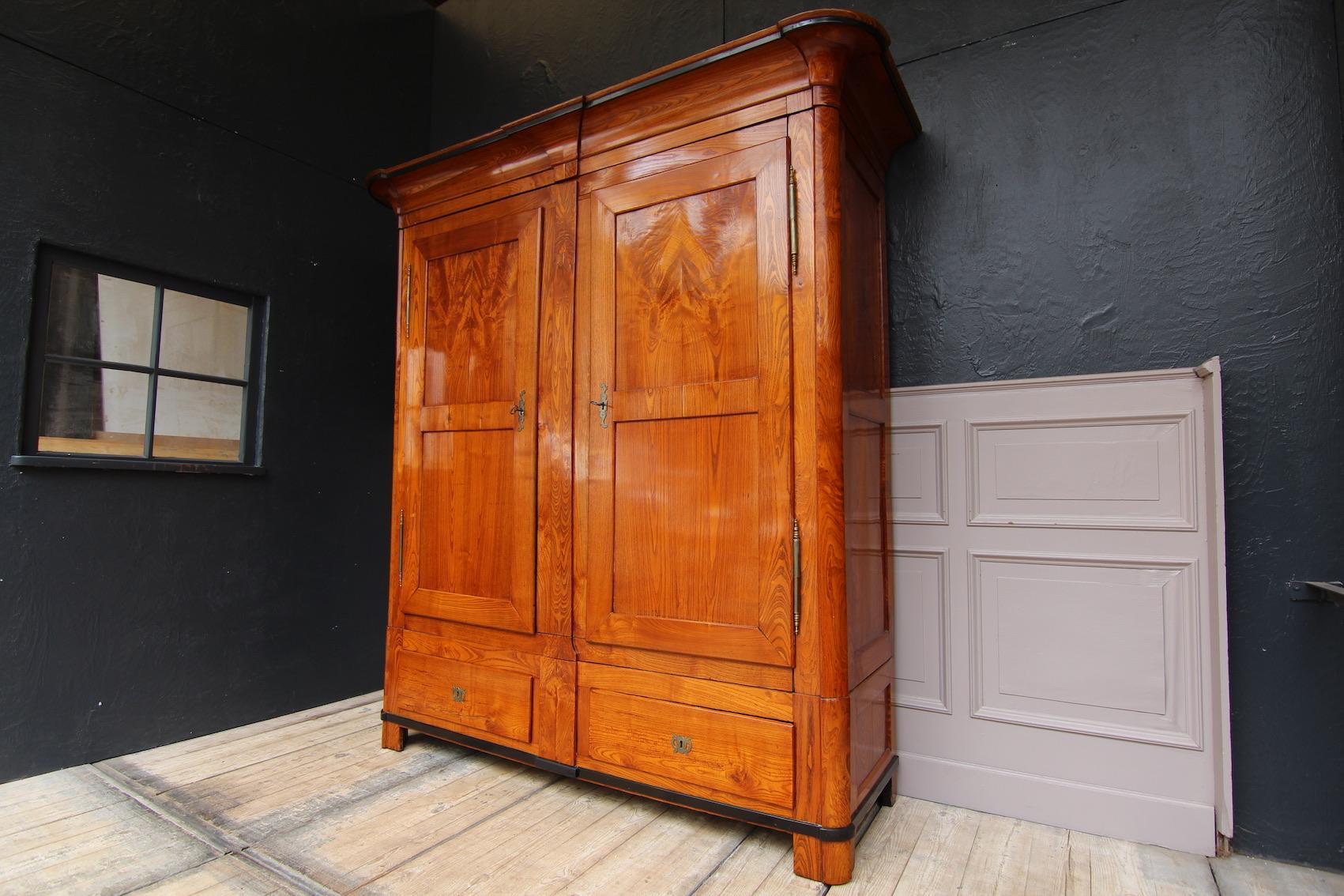 Biedermeier wardrobe made of ash wood. 1st half of the 19th century.
2-door coffered body with cranked rounded corners and head strip with center console. Base with 2 drawers and a secret compartment in the middle (see photos). Both the cornice and