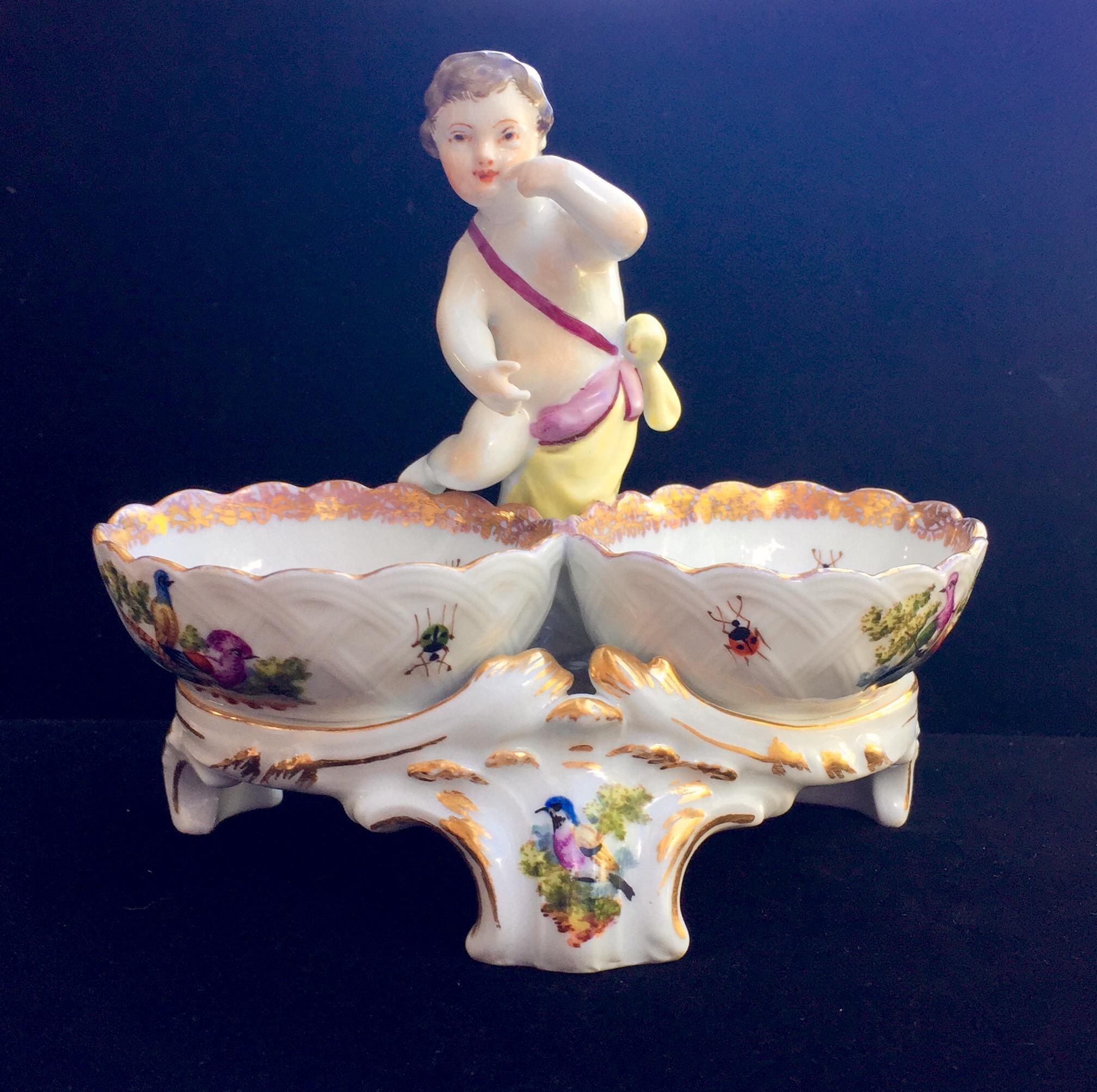 This open salt was made in the 19th century in typical Rococo style. The interior of the salt basin and the stand is delicately painted with birds, flowers and insects. In the center, on top, stands a figure of an gesticulating cherub. 

Delicate