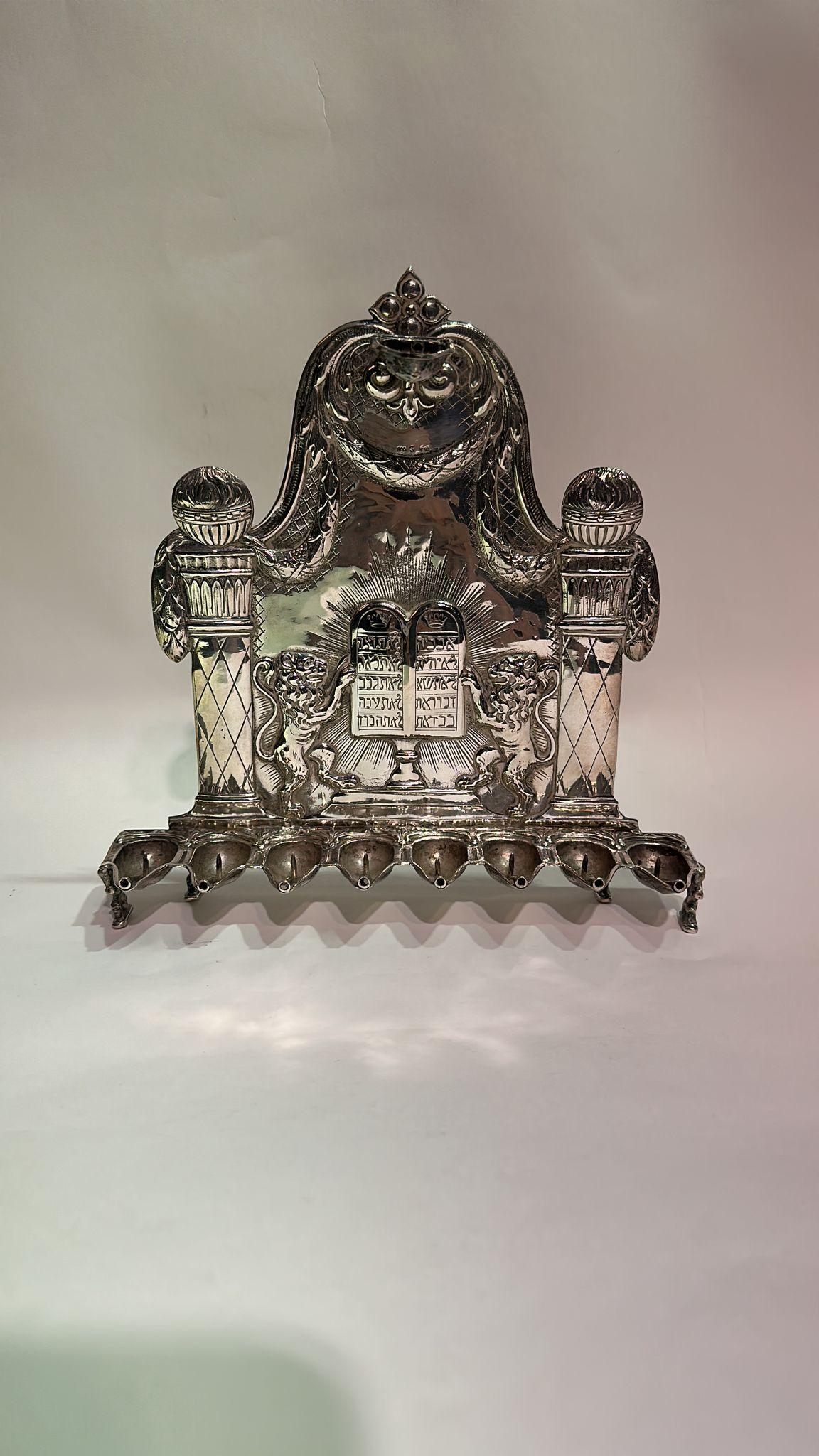 Si tratta di una menorah di Hanukkah in argento tedesca (hanukkiah), che riflette l'abilità artigianale e le tradizioni artistiche degli argentieri tedeschi del XVIII o XIX secolo.
Nel pannello centrale: I Dieci Comandamenti, incisi in ebraico, sono