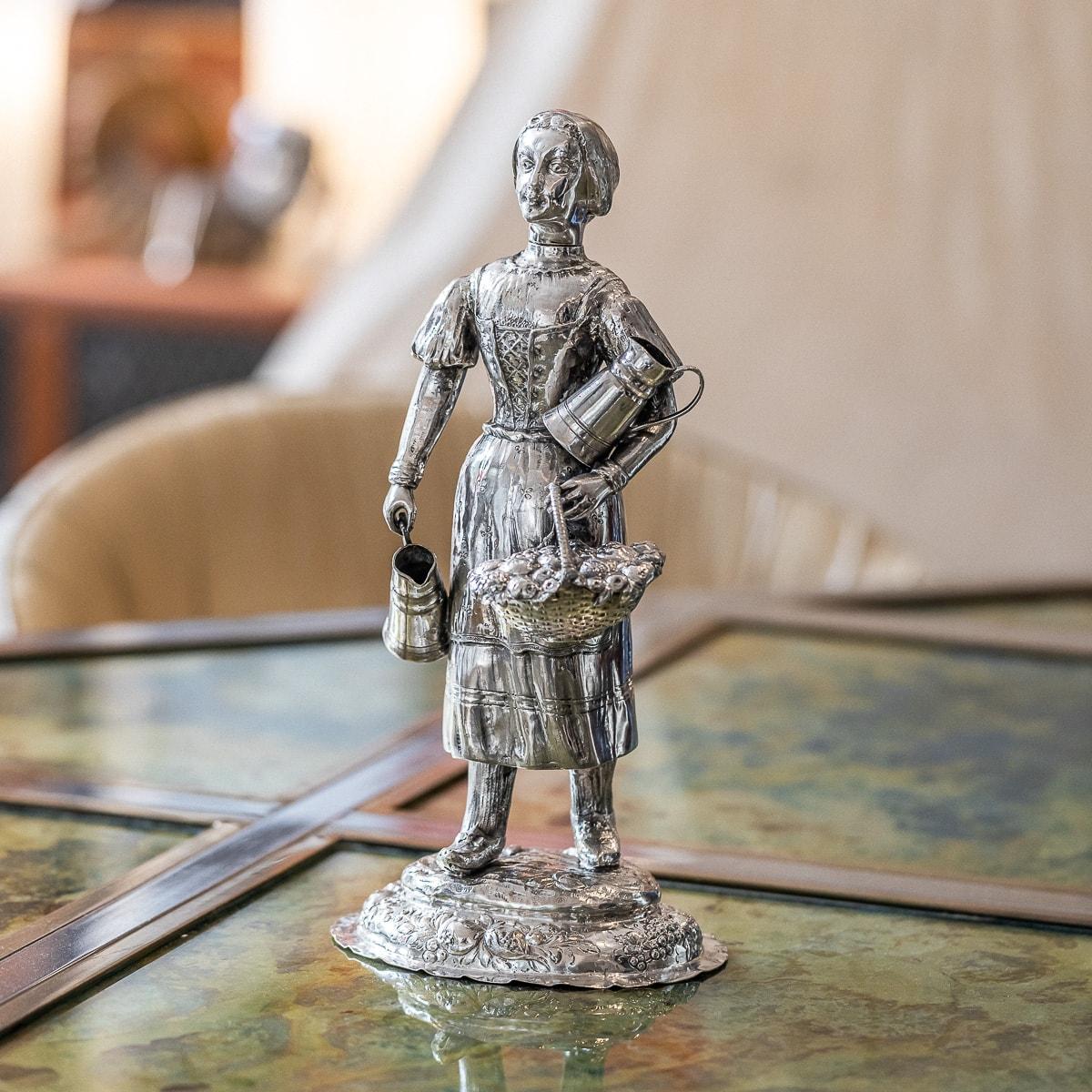 Antique 19th Century German solid silver figural table ornament modelled as a female fruit seller, standing on a large shaped-oval base embossed with flowers, right hand holding a water pitcher and one resting on the left, the left hand holding a