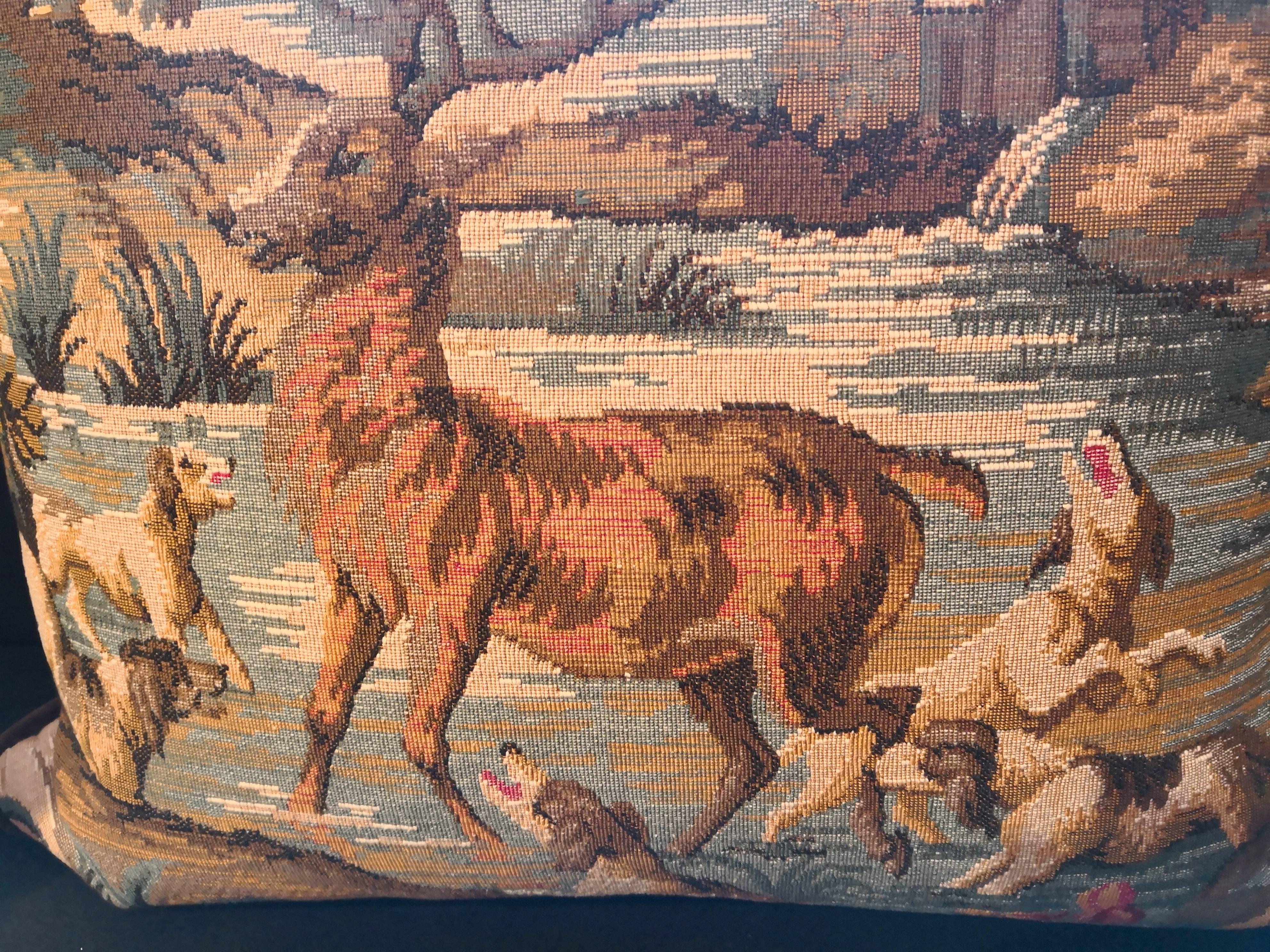 19th century German tapestry fragment made into a cushion. Backside dark brown velvet. The tapestry shows a traditional hunting scene with a dear and dogs. In the background a landscape in blue and yellow. Framed with dark brown velvet and backside