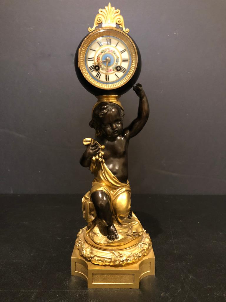 pendule figurative de style Louis XVI du XIXe siècle. Putti doré et patiné sur socle en bronze supportant une horloge en forme de boule avec cadran en porcelaine.