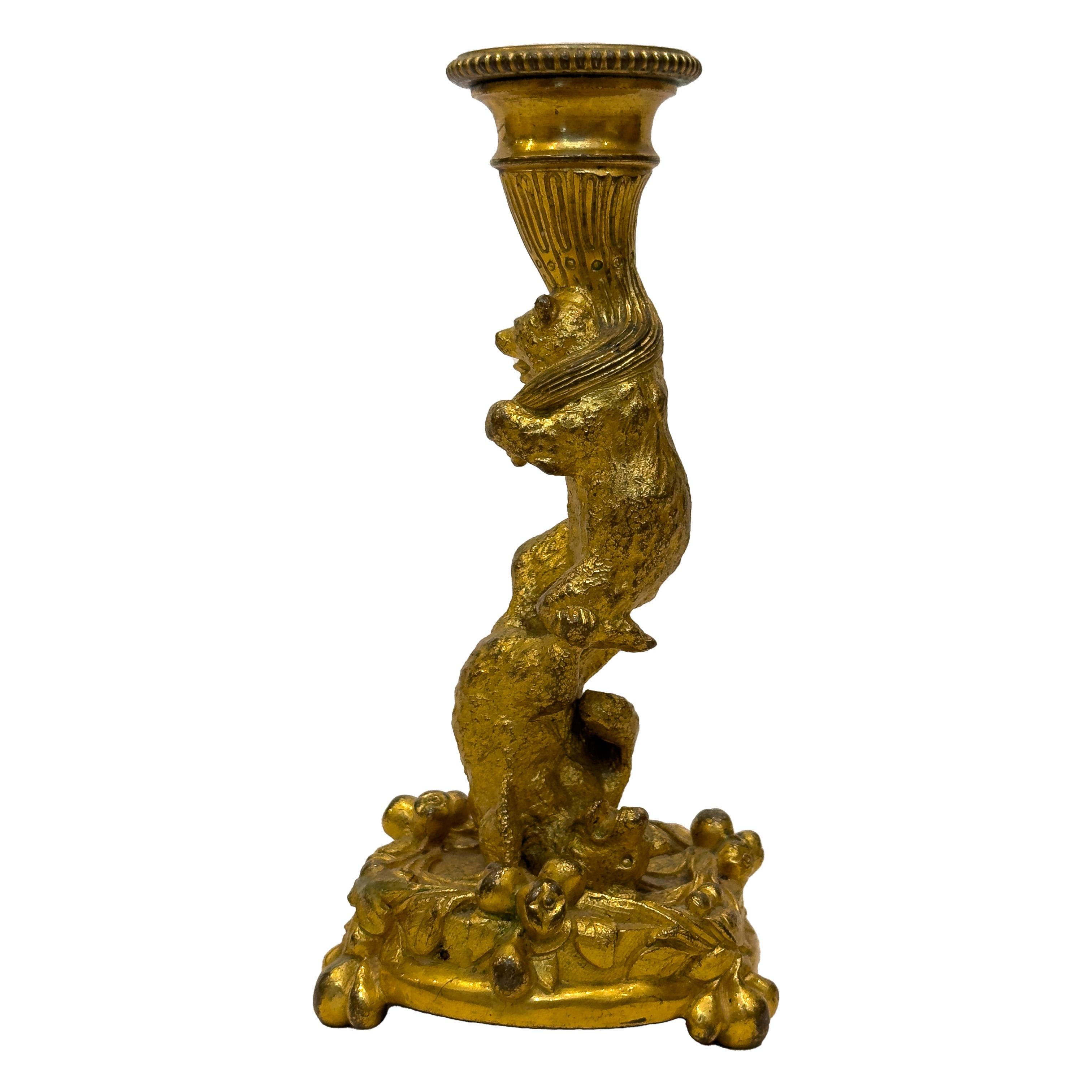 Un candeliere in bronzo dorato con un orso acrobatico che tiene in mano una betta attorcigliata e ornata di canestrini. Su una base rotonda a forma di fiore, l'orso sostiene il portacandele. Il candeliere poggia su una base circolare a tripode