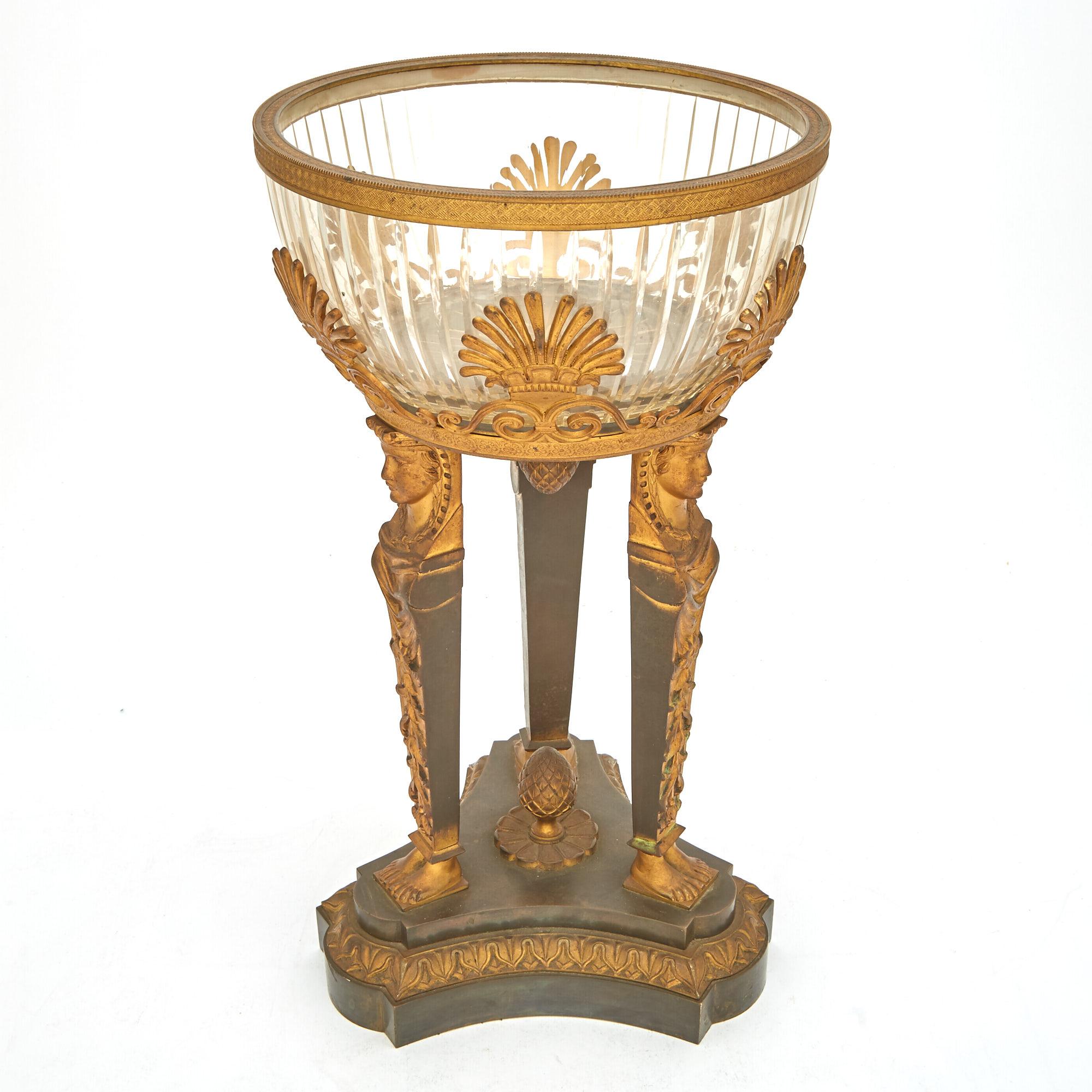 Adéntrate en el esplendor del siglo XIX con este excepcional centro de mesa de estilo Imperio, una cautivadora unión de monturas de bronce dorado y cristal tallado que personifica la opulencia de la época. En su centro hay un recipiente circular