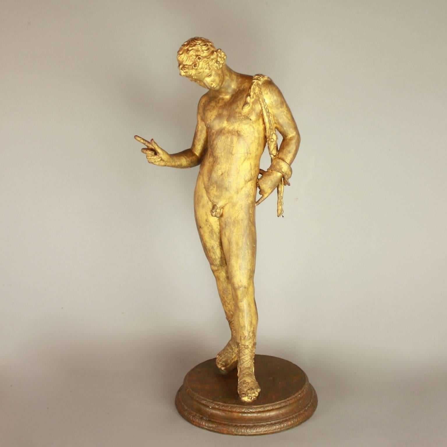 The gilt-bronze statue of a nude youthful Dionysus standing on his right leg. In contrapposto position sculpted with a well-defined musculature. The head is bent down with a contemplative gaze. He wears a crown of ivy and has a fawn-skin knotted