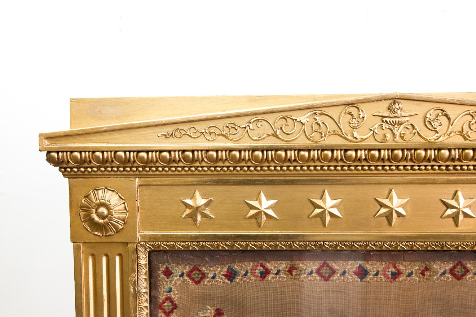 19th century gilt needlework fire screen, the crest having gilt metal urn and flame with trailing arabesques, the frieze with star appliques, reeded stiles with applied rosettes on each corner, floral needlework behind glass, stylized leaves applied