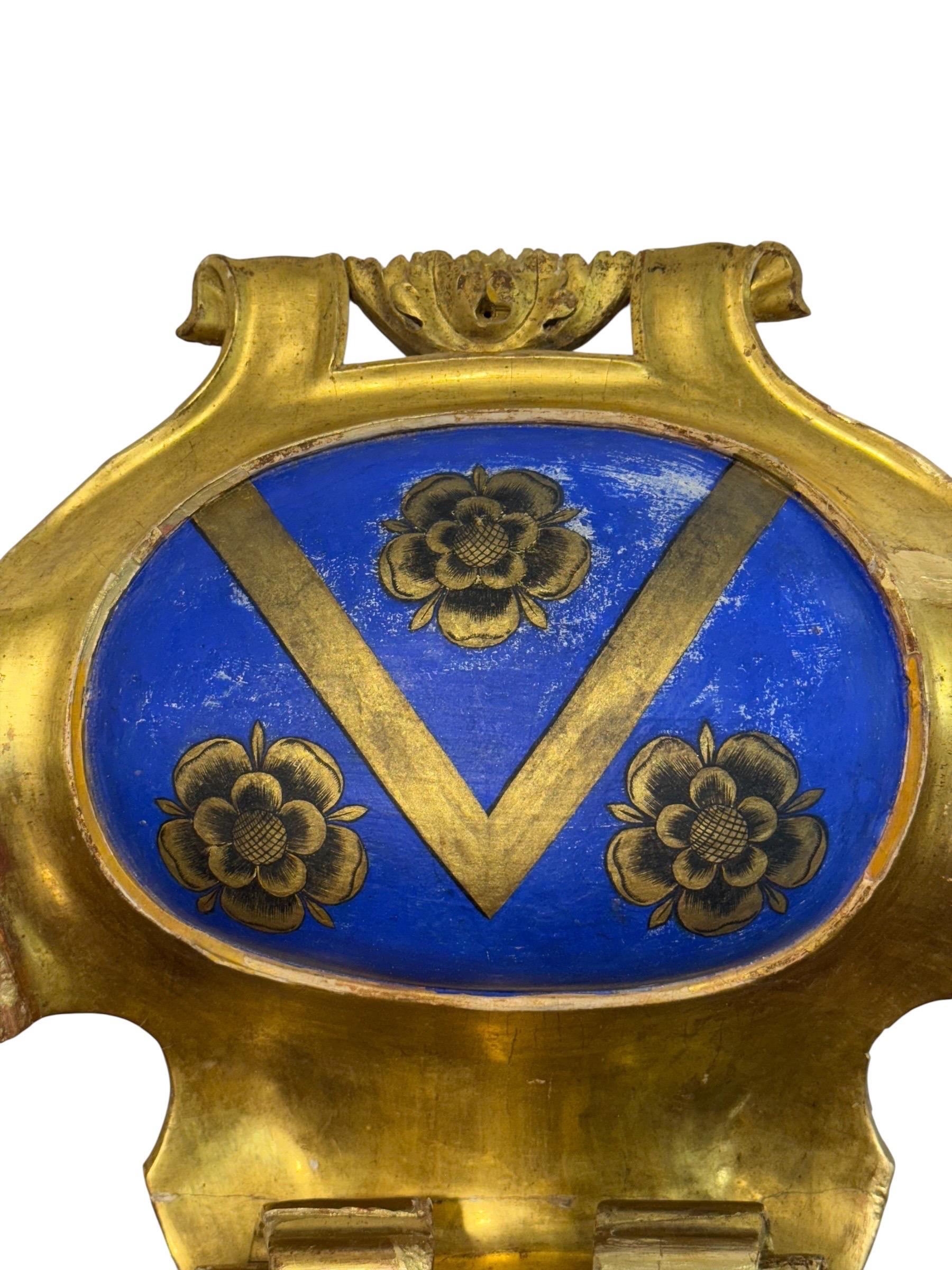 Uno scudo in legno scolpito, gesso, dorato ad acqua e policromato, risalente al XIX secolo. Il cartiglio sagomato incornicia una riserva armoriale ovale dipinta in un saturo blu cobalto (azzurro). Al centro c'è un ampio chevron a forma di V in