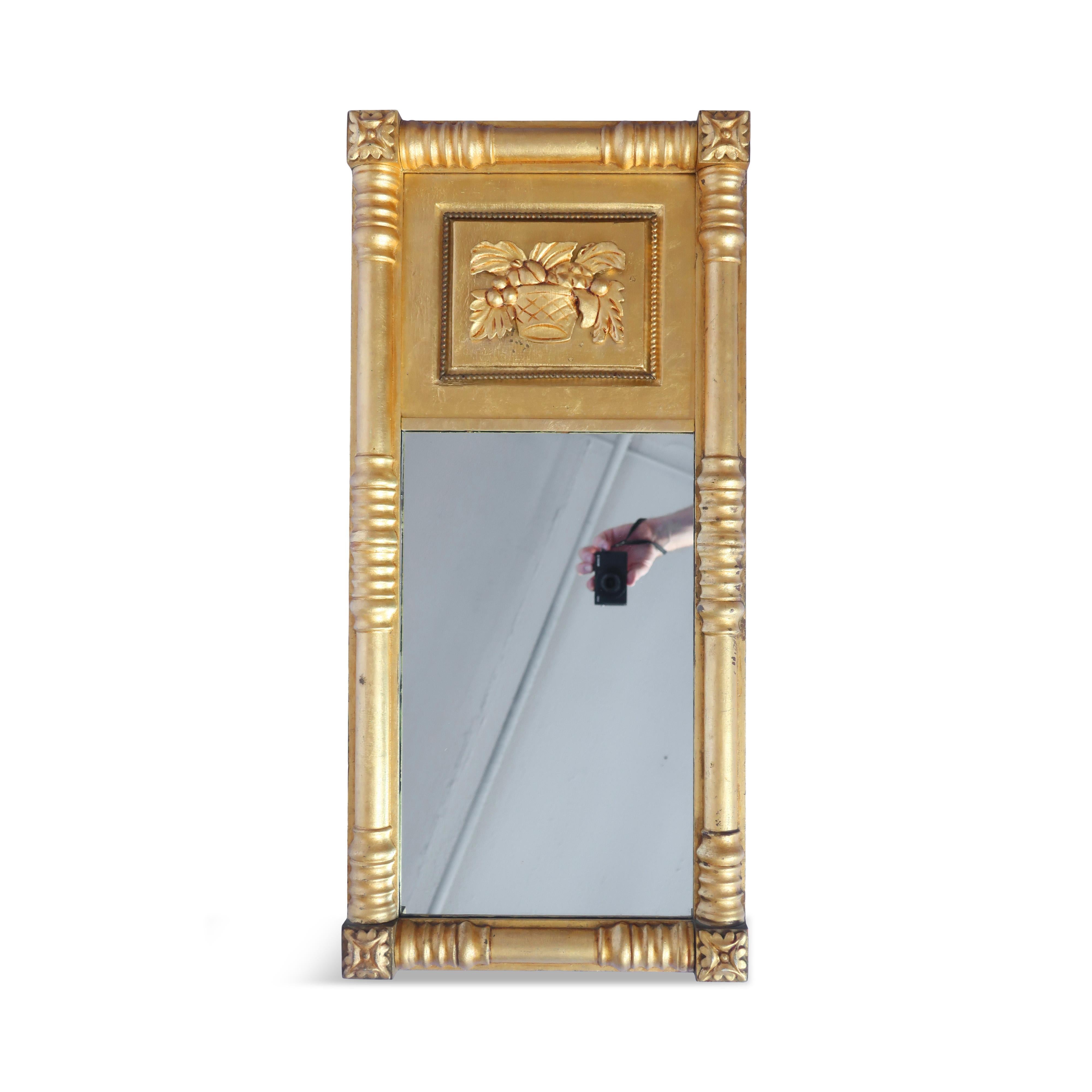 An antique carved giltwood wall mirror from the 1800s featuring a rectangular frame with rosette corners and a basket of fruit panel at the top.  It is made of wood and gilded with gold leaf, giving it a rich and elegant look; it is suitable for