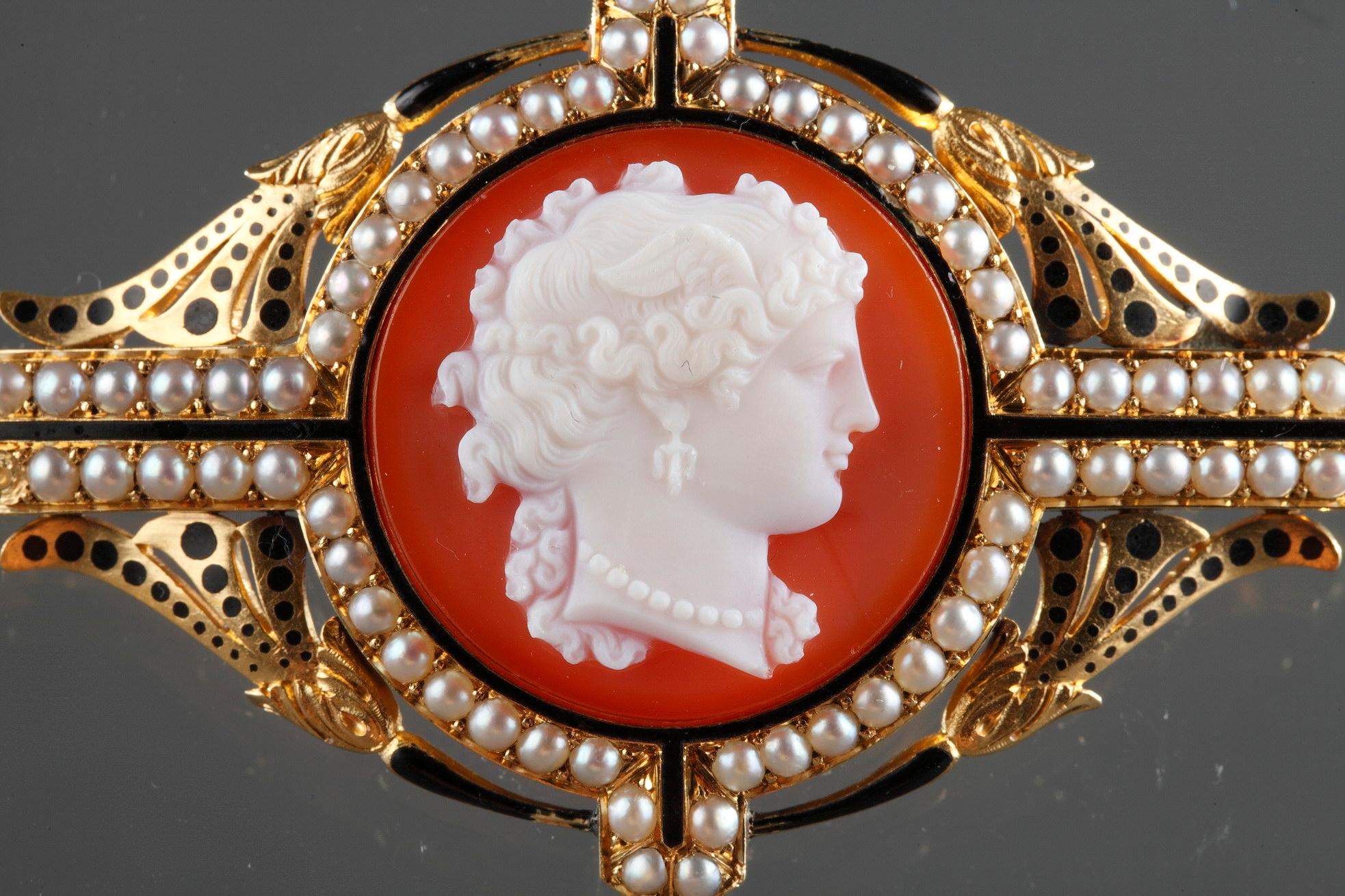 Spilla a cammeo in agata montata in oro che raffigura il busto di una giovane donna con una collana di perle. Montature in oro traforato decorate con perle e motivi floreali in smalto nero. Chiusura antica.
Punzone francese per l'oro: testa d'aquila