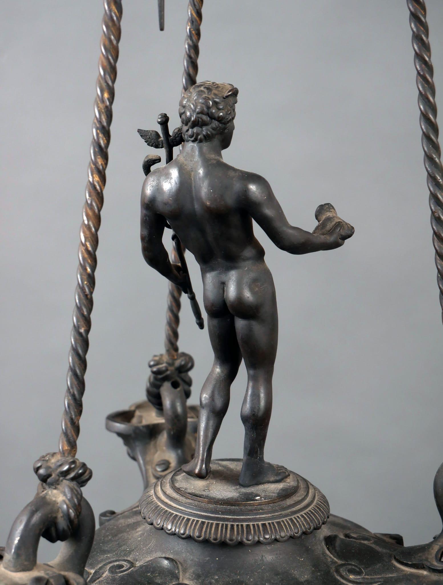 Questa lampada ad olio a sospensione romana, decorativa e di grande effetto, risalente all'inizio del XIX secolo, è una copia di un originale ritrovato a Pompei. Mercurio, il dio del commercio, dei ladri e dei viaggiatori, è raffigurato al centro