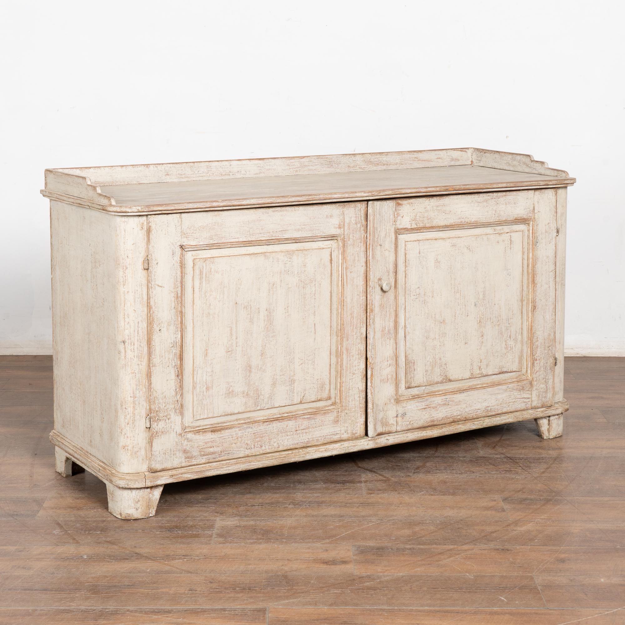 Credenza o mobile server Gustavo in pino verniciato grigio chiaro con grandi ante e ripiano interno singolo.
La finitura verniciata di recente, applicata professionalmente e di colore grigio chiaro, è leggermente consumata per adattarsi all'età e