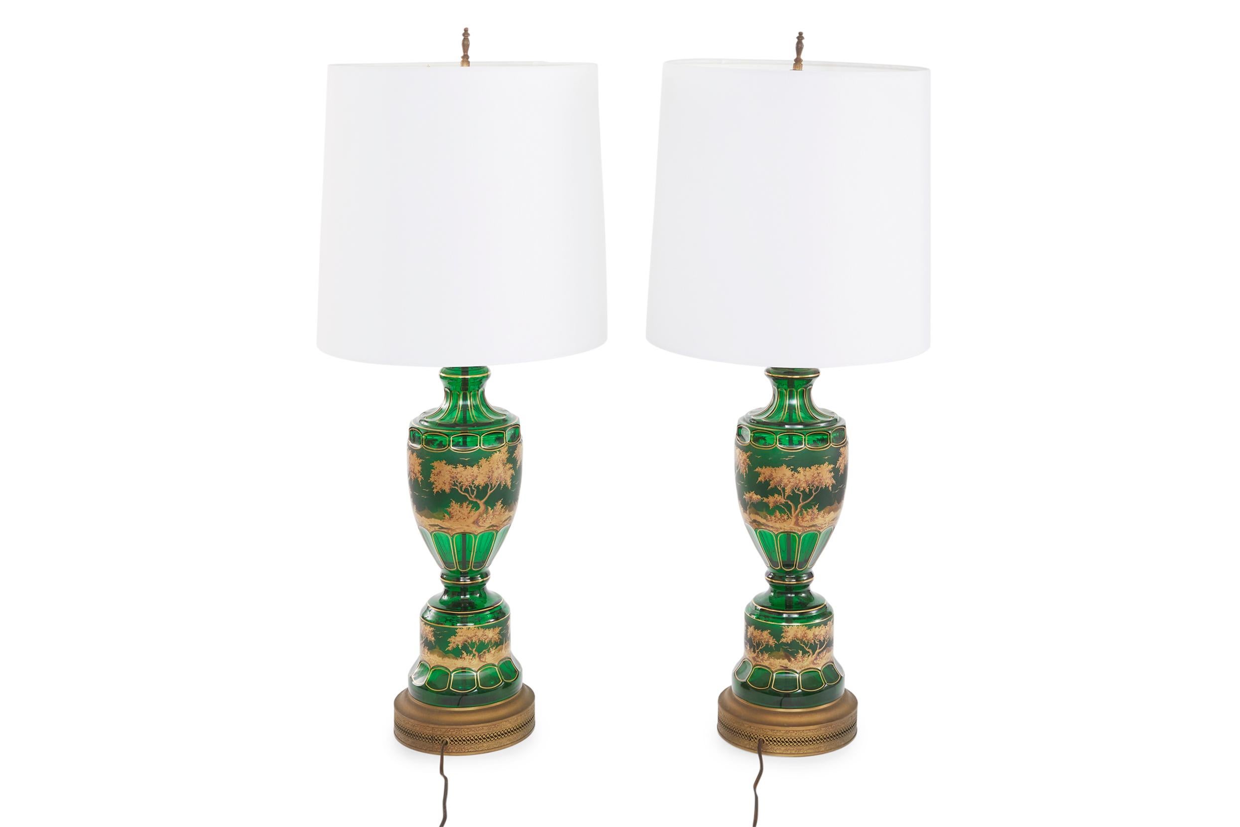 19th century green with gold scene design details Moser glass pair table lamp with brass base. Each lamp is in good working condition. Minor wear consistent with age / use. Each lamp stands about 35.5 inches X 8 inches. Each drum shade is 13 inches