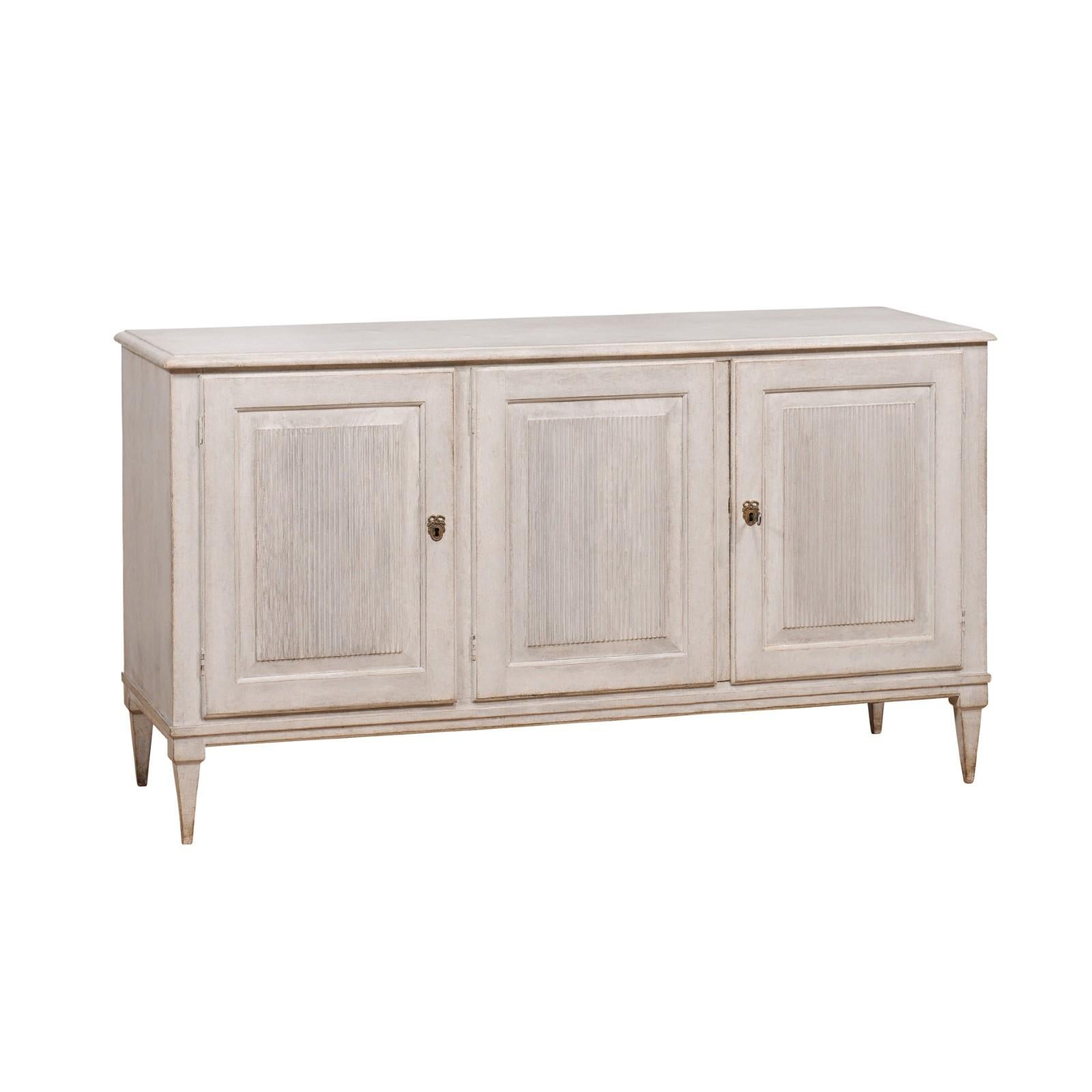 A Swedish Gustavian style sideboard from the 19th century with antique light gray finish, three carved reeded doors, inner drawer and tapered legs. Channeling the timeless beauty of Swedish craftsmanship, this Gustavian style sideboard from the 19th