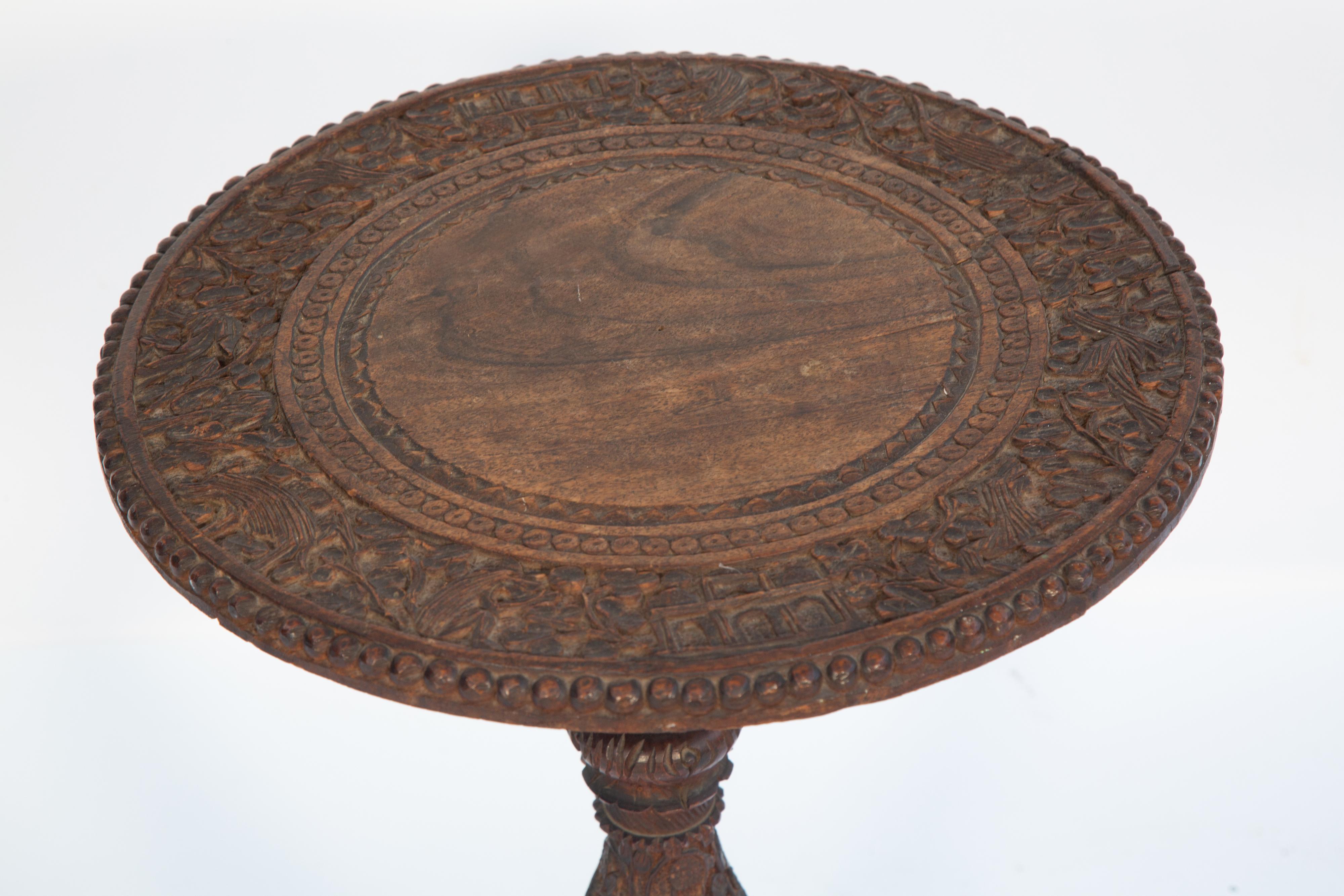 Table d'appoint à chandelier, en bois de teck sculpté de manière élaborée, avec un plateau rond sur une base piédestale très décorée, reposant sur des pieds évasés se terminant par un trio de dauphins, sur des pieds en coussinets. 

Diamètre de la