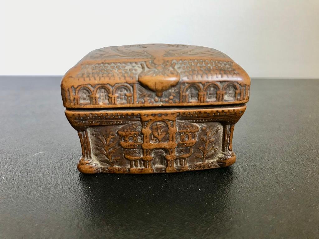 A wonderfully carved Coquilla nut tobacco or snuff box. Portuguese, first half of the 19th century. The top with a portrait of woman holding a flower with a ribbon in her hair. The front an interesting architectural fantasy of an edifice with