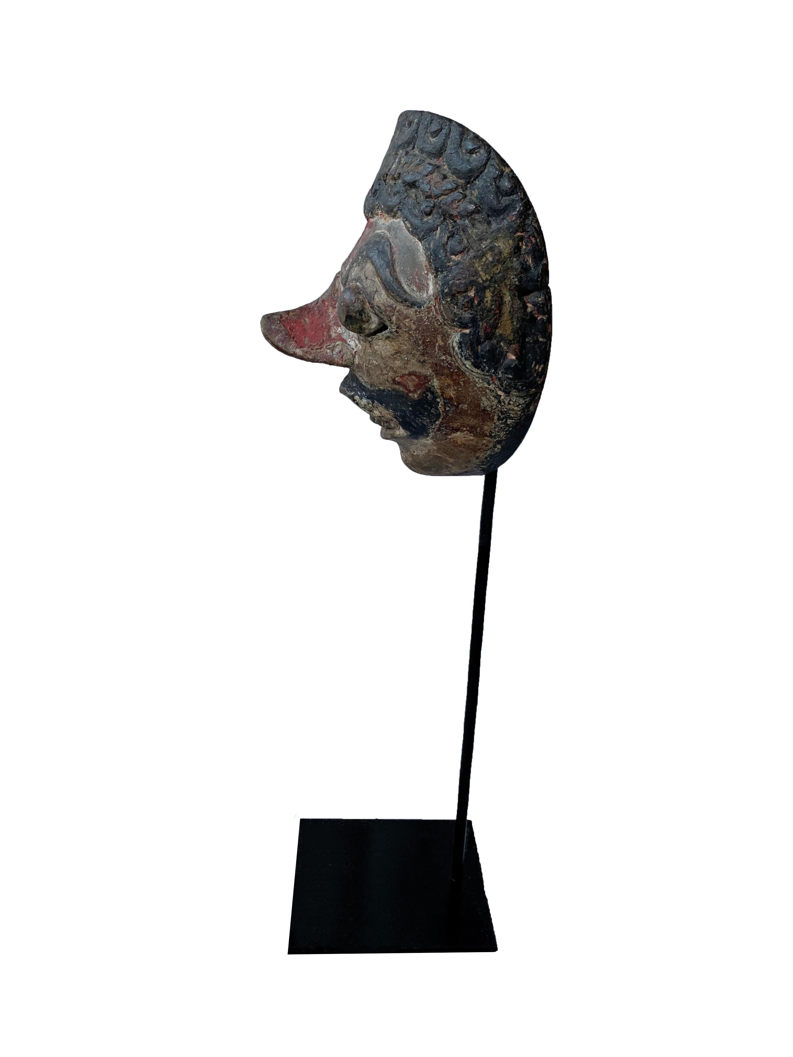 A visibly old mask from the Javanese masks theatre ‘wayang topeng’: carved from hard wood, and painted on the front side. This mask is from Cirebon in Java. With dark, reddish skin and slits either side of its pupils for the eyes through which the