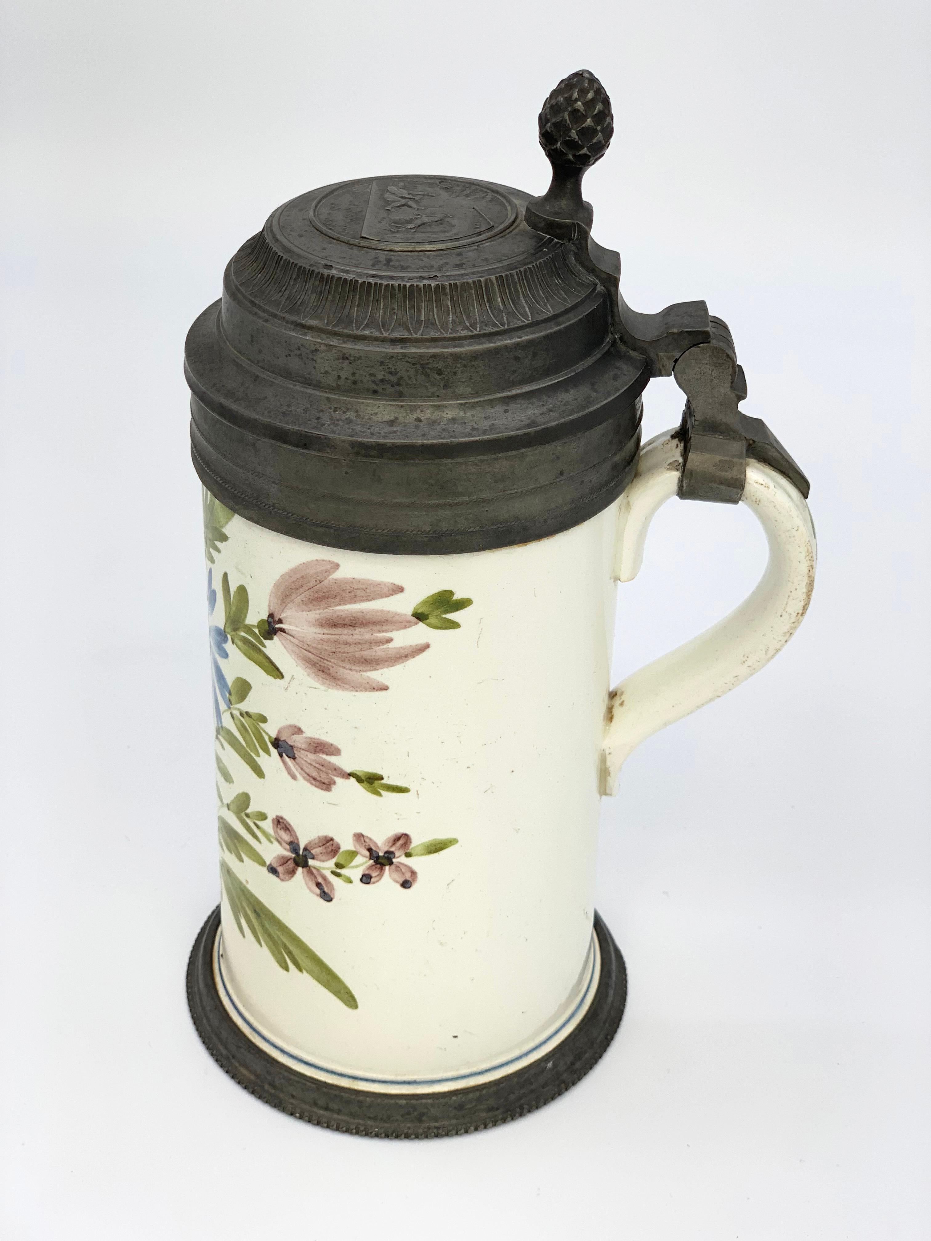 Beautiful 19th century beer mug with hand painted flowers in blue, purple and green.
Tin bottom and lid with an engraved scene with a drunkard and a bock.

     